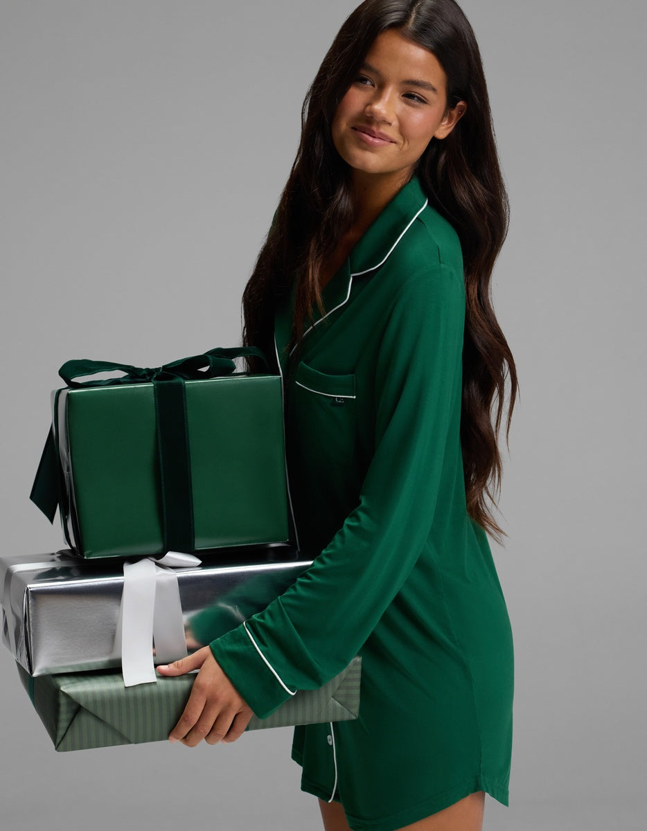 A woman wearing a dark green Cozy Earth Women’s Bamboo Stretch-Knit Long Sleeve Sleep Dress with white piping holds a stack of three gift-wrapped boxes.