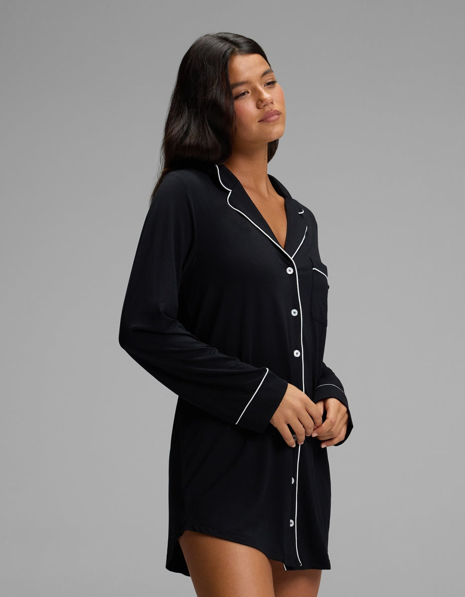 A person wears a black Cozy Earth Women’s Bamboo Stretch-Knit Long Sleeve Sleep Dress featuring white piping detail.