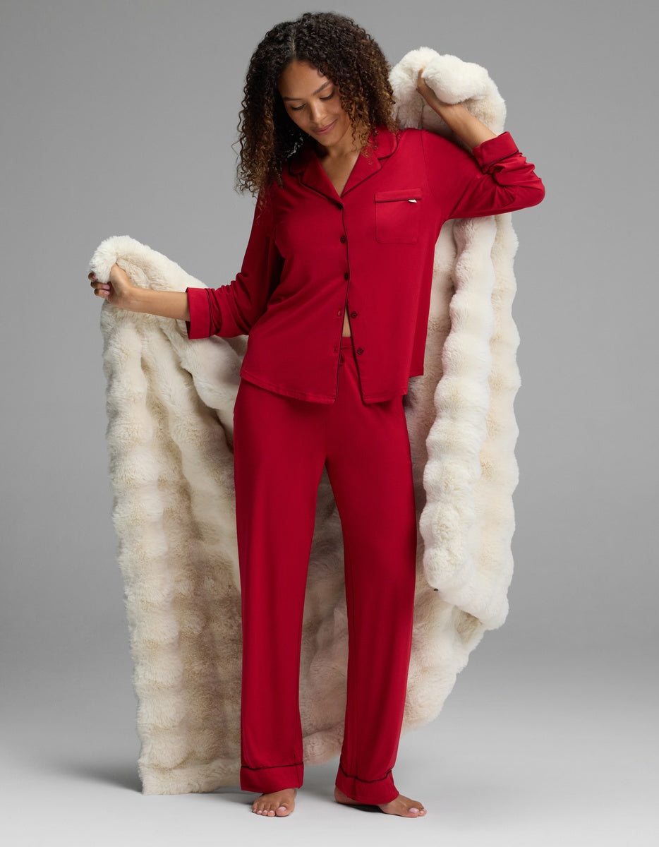 A woman wearing a red button-down pajama top and matching Women’s Bamboo Stretch-Knit Pajama Pant, wrapped in a large, cream-colored plush blanket.