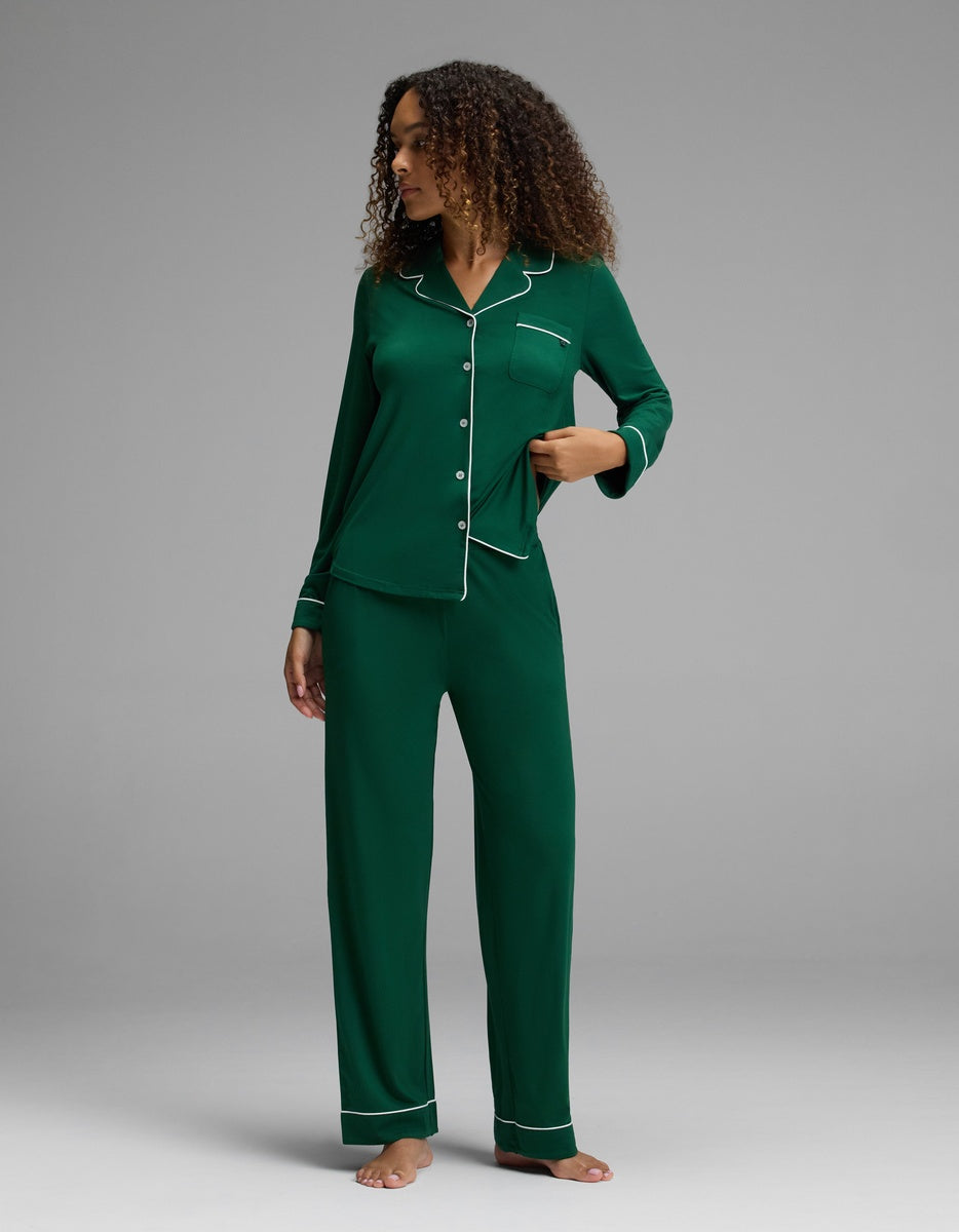 A person wearing a dark green Women’s Bamboo Stretch-Knit Pajama Pant set with white piping on the button-up top and long pants.