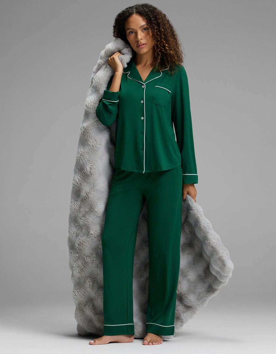 A woman wearing dark green Women’s Bamboo Stretch-Knit Pajama Pant and a matching button-down top with white piping, draped in a textured grey blanket.