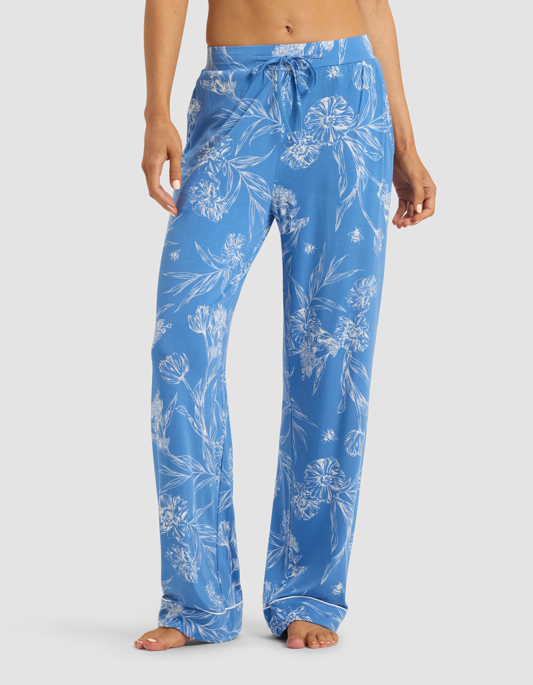 A woman models the Cozy Earth Women's Bamboo Stretch-Knit Pajama Pant, featuring a blue fabric with a white floral pattern. Barefoot, she stands against a plain background, showing only her lower torso and legs.