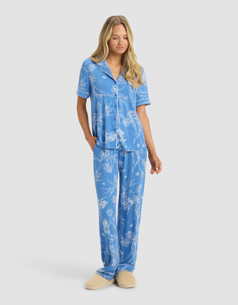 A woman with long blonde hair wears Cozy Earth's Women's Bamboo Stretch-Knit Pajama Pant in blue with a white floral pattern and beige slippers, standing forward with one hand in her pocket against a plain light background.