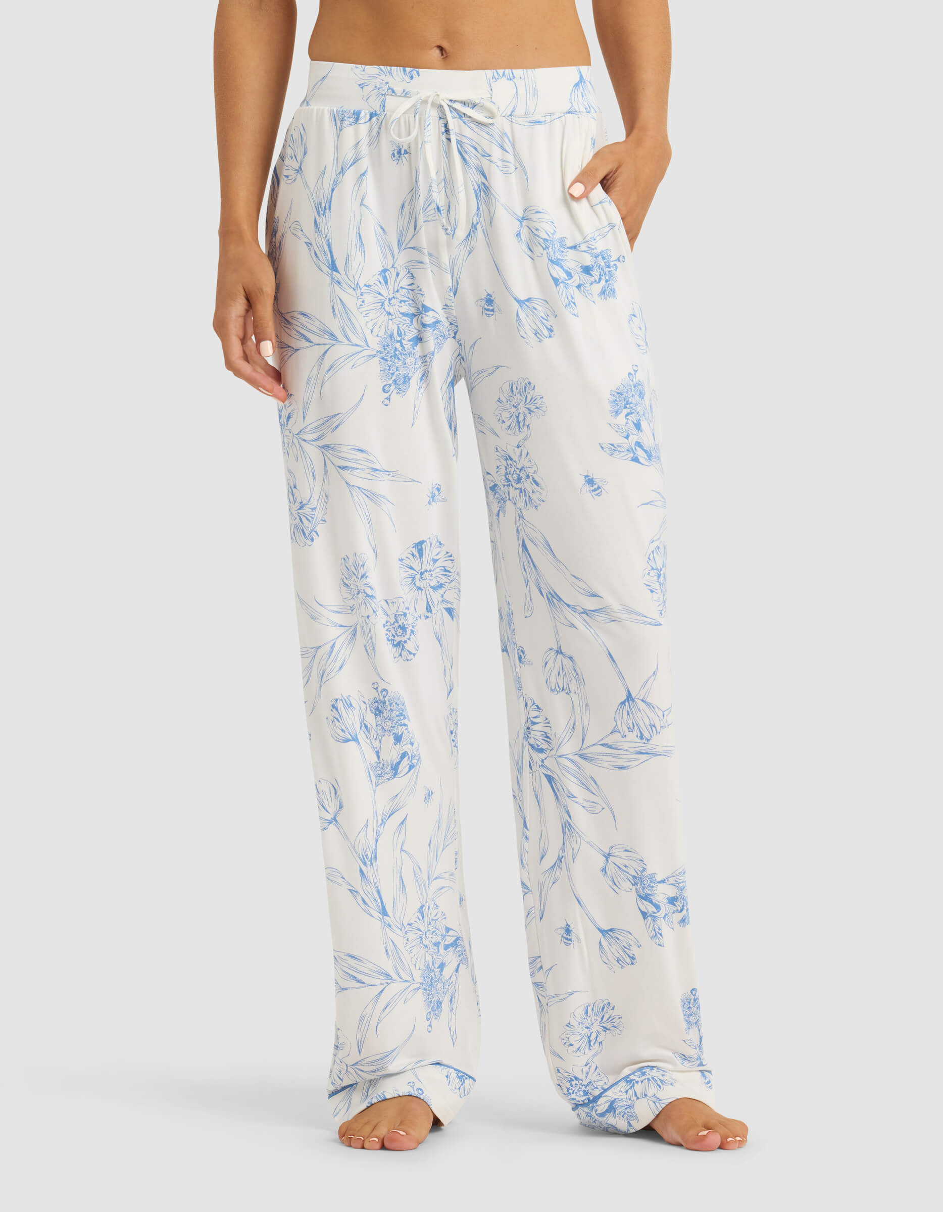 A woman wears Cozy Earth's Women's Bamboo Stretch-Knit Pajama Pant—loose, white with blue floral print and a drawstring waist—standing barefoot with hands in pockets against a plain background.|Color:Garden Toile in Sea Salt