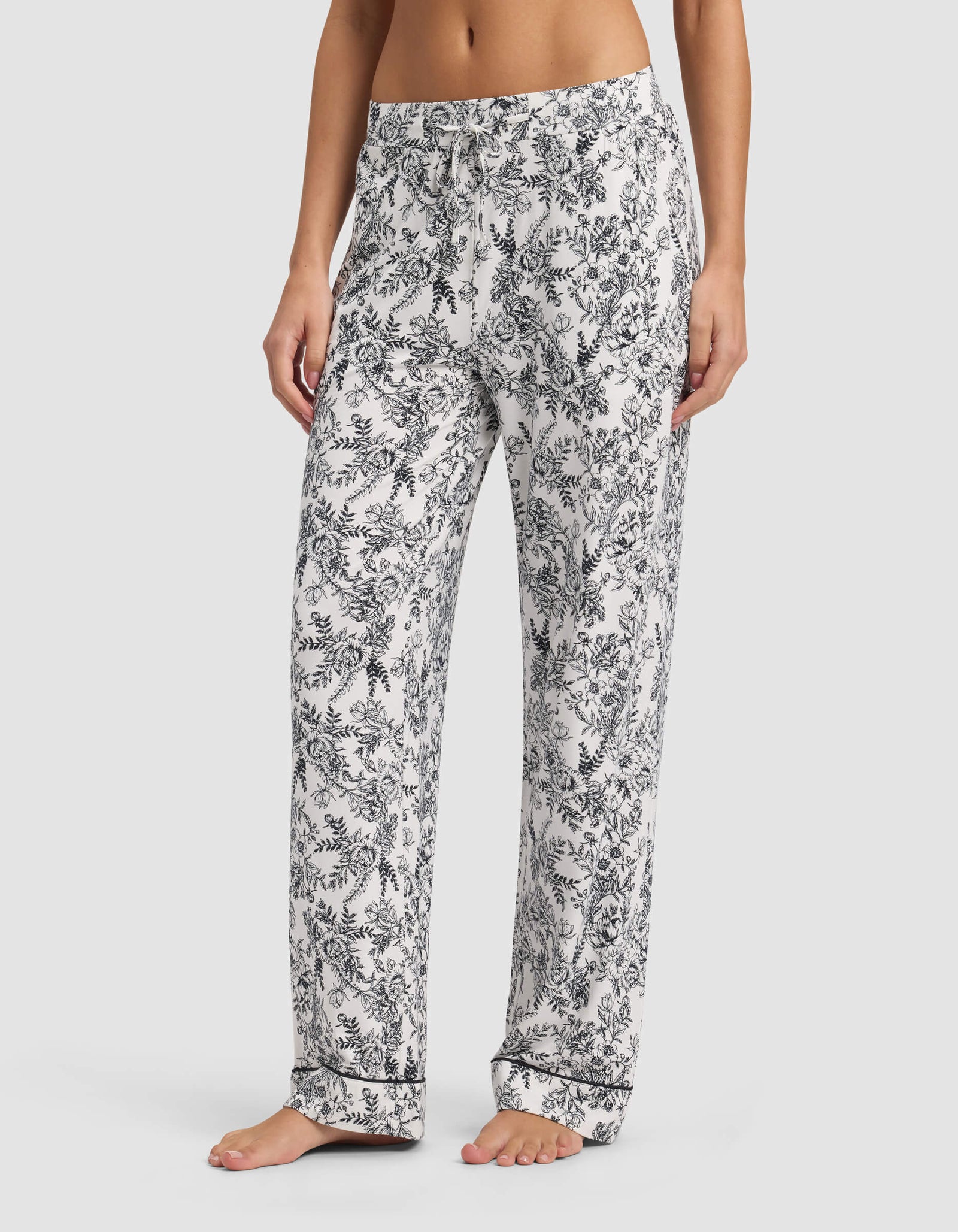 A woman stands barefoot wearing Cozy Earth’s Women's Bamboo Stretch-Knit Long Sleeve Pajama Set in a loose fit with a black floral pattern and drawstring waist. The image is cropped from her midriff to her feet.