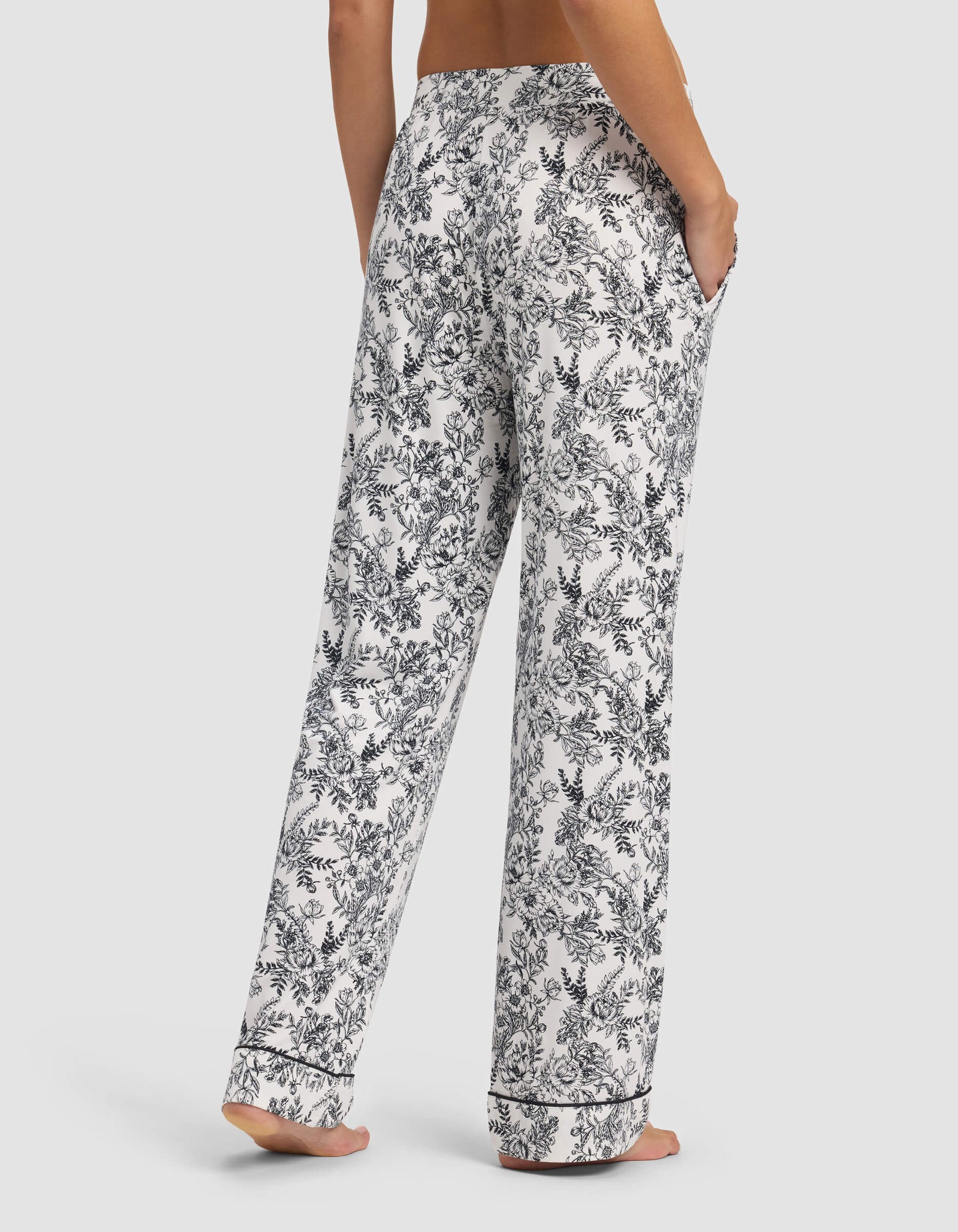 A person stands barefoot, shown from the waist down, wearing Cozy Earth Women's Bamboo Stretch-Knit Pajama Pants featuring a black floral pattern. Hands are in pockets and the backdrop is a plain, light color.