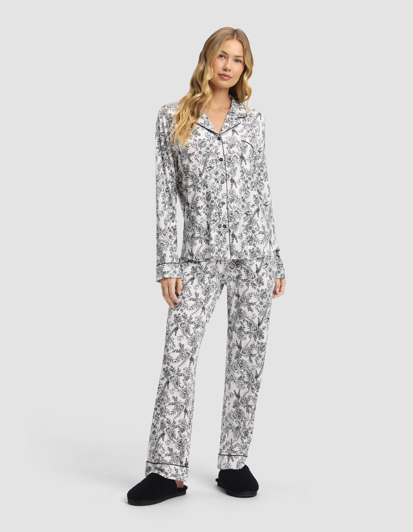 A woman poses against a light gray background wearing Cozy Earth Women's Bamboo Stretch-Knit Pajama Pants in a black and white floral pattern, paired with matching long-sleeve top and black slippers.