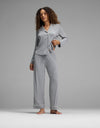A woman with curly hair stands barefoot against a gray backdrop, wearing the Cozy Earth Women’s Bamboo Stretch-Knit Long Sleeve Pajama Set - Petite in light gray. She is holding up the hem of her pajama top slightly.|Color:Light Heather Grey