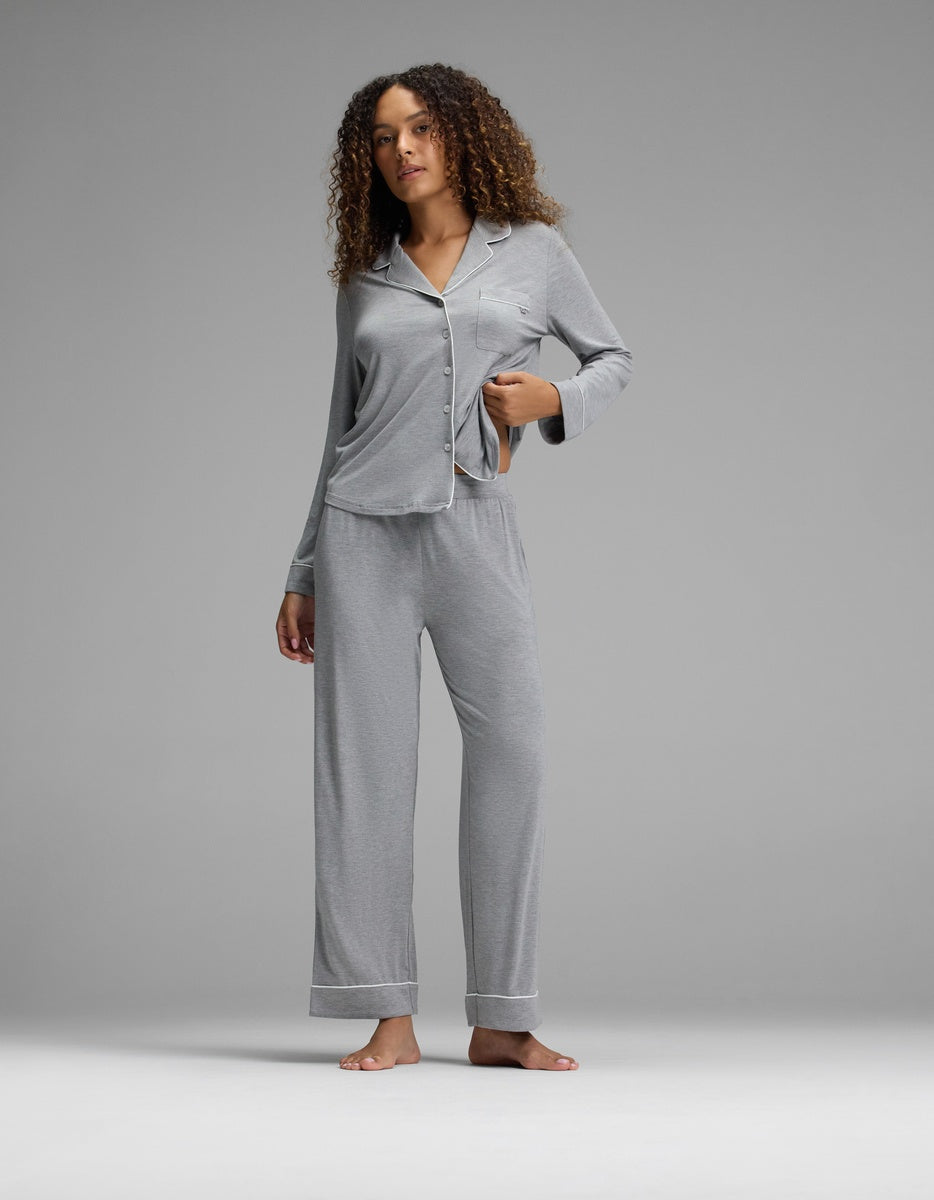 A woman with curly hair stands barefoot against a gray backdrop, wearing the Cozy Earth Women’s Bamboo Stretch-Knit Long Sleeve Pajama Set - Petite in light gray. She is holding up the hem of her pajama top slightly.