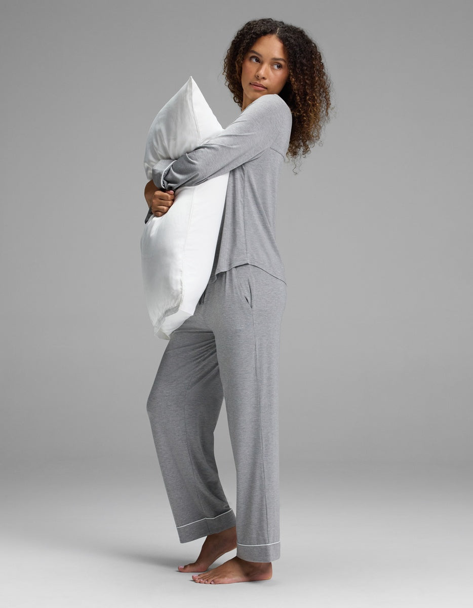 A woman with curly hair stands barefoot on a plain backdrop, holding a large white pillow against her chest. She wears the Cozy Earth Women’s Bamboo Stretch-Knit Long Sleeve Pajama Set - Petite in gray and looks slightly to the side with a neutral expression.