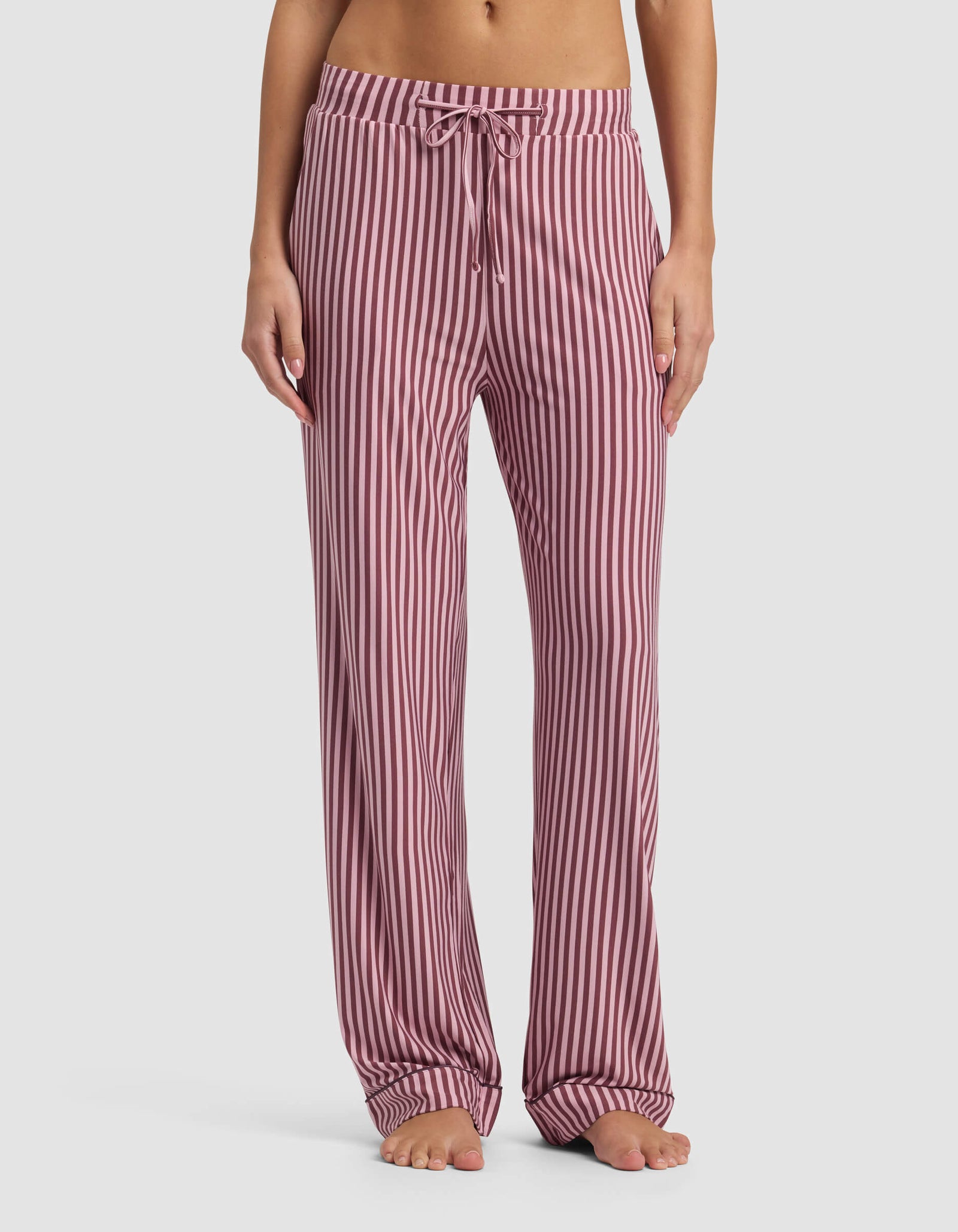 A person stands barefoot, showing only the lower torso and legs, wearing Cozy Earth Women's Bamboo Stretch-Knit Pajama Pant in pink and white vertical stripes with a drawstring waist against a plain background.