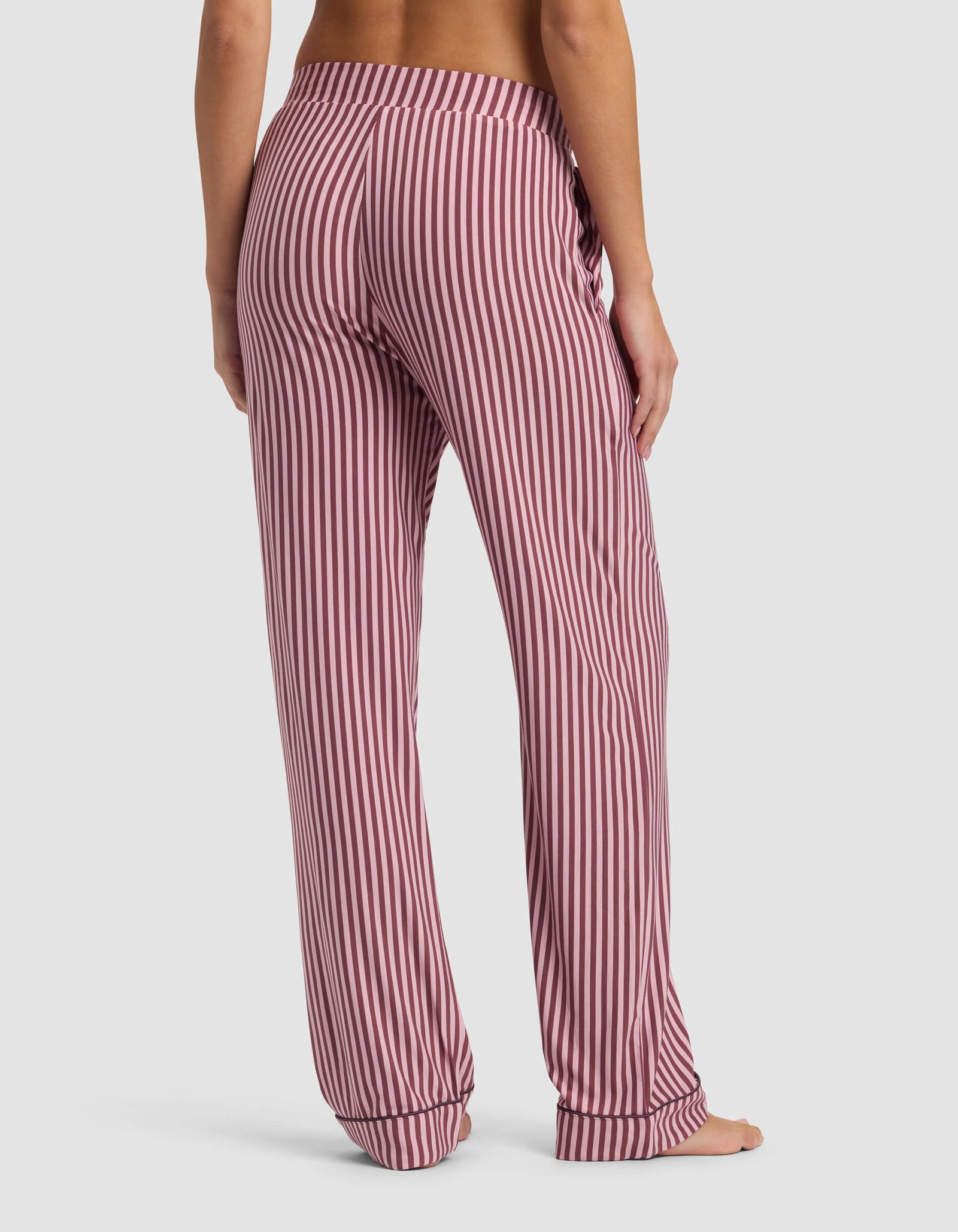 A woman stands barefoot on a white background, shown from the back, wearing Cozy Earth's Women's Bamboo Stretch-Knit Pajama Pant with pink-and-white vertical stripes, an elastic waistband, and a relaxed fit.
