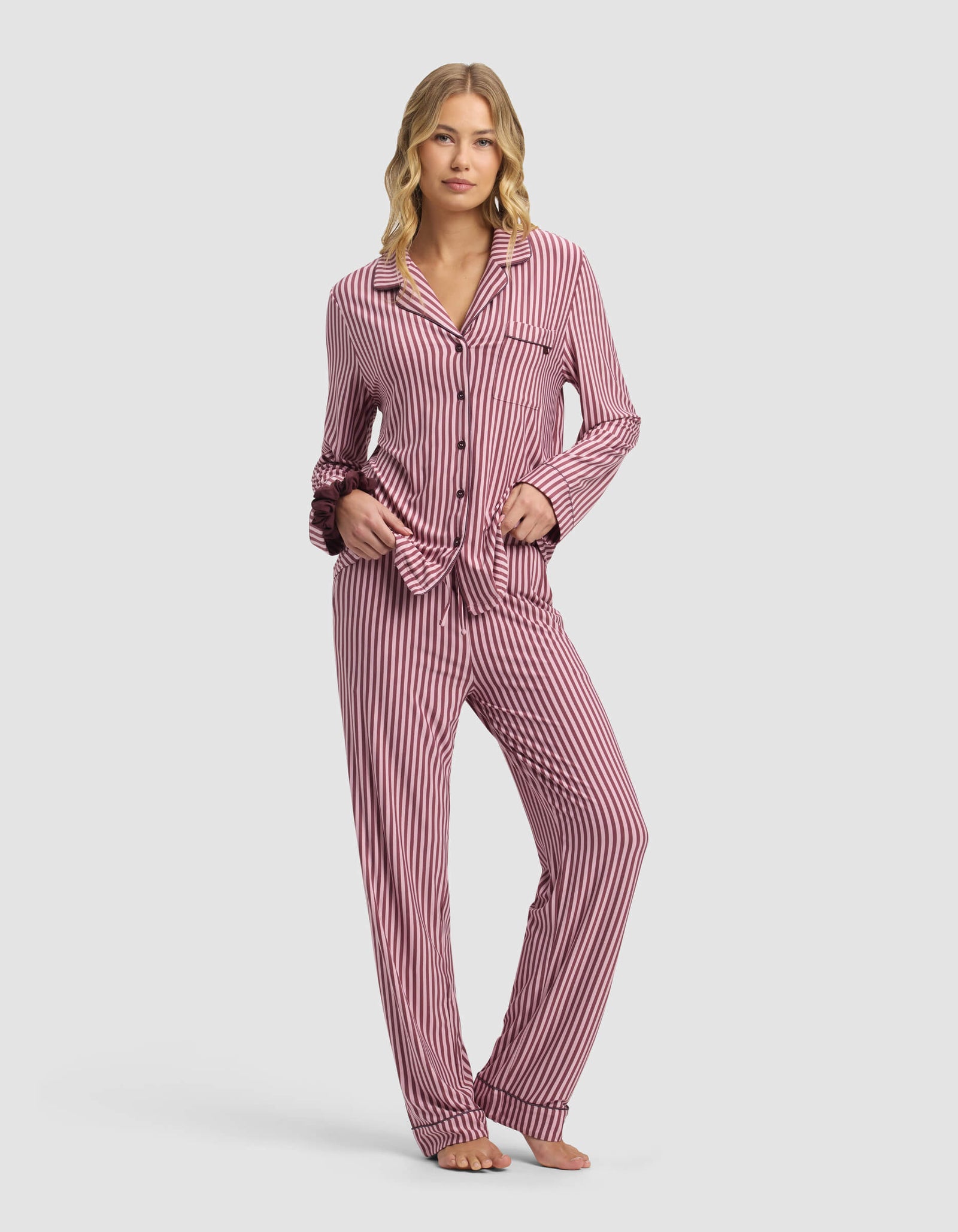 A woman stands barefoot wearing Cozy Earth Women's Bamboo Stretch-Knit Pajama Pant in pink and white vertical stripes, with long sleeves, buttons down the front, one hand in her pocket and the other on her tie-front top.