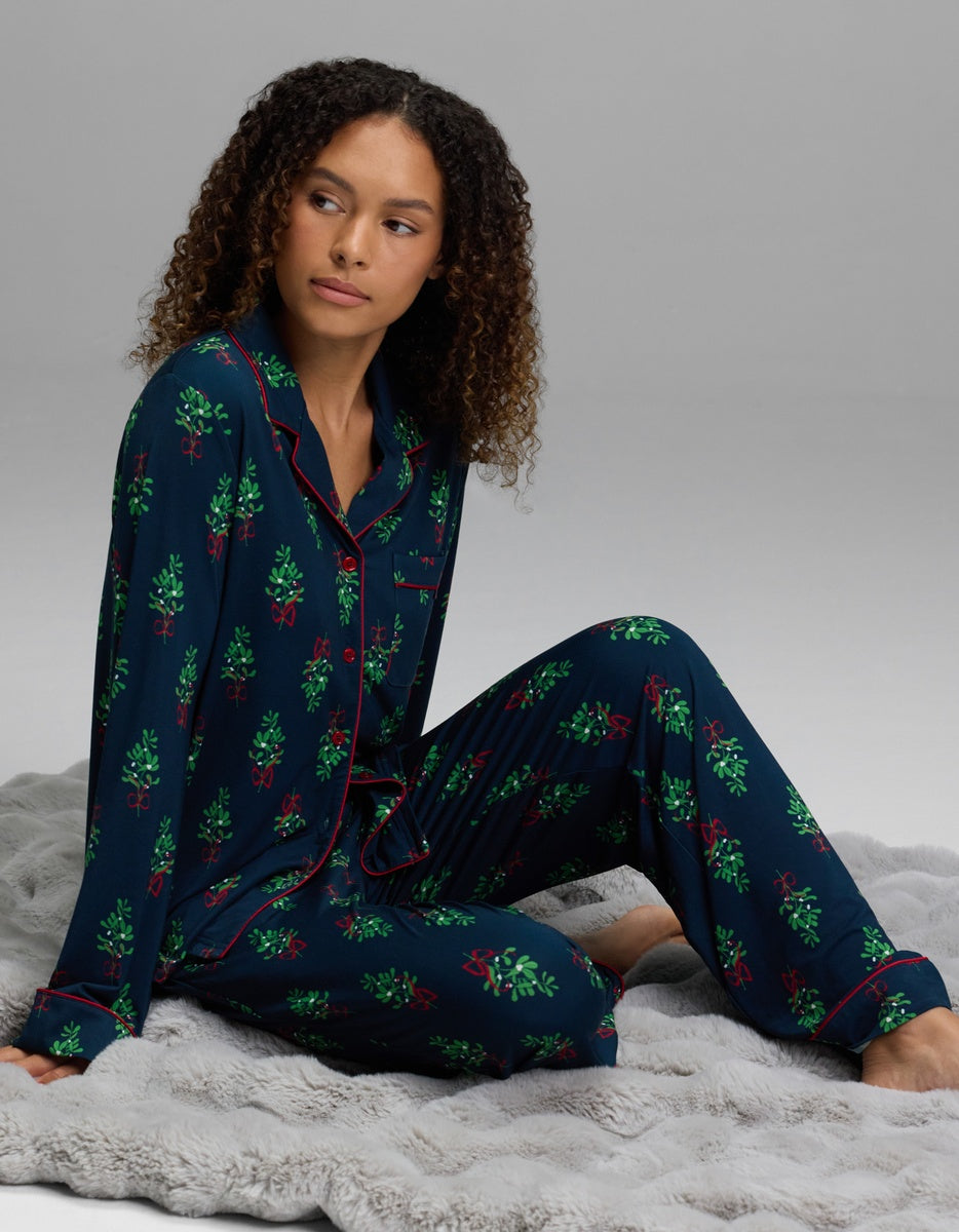A woman wearing dark blue long-sleeved pajamas patterned with mistletoe and red piping, including Women’s Bamboo Stretch-Knit Pajama Pant.