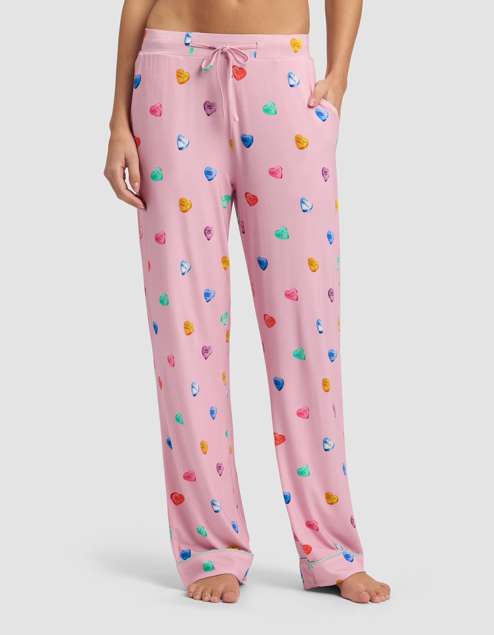 A person stands barefoot, showing only their lower torso and legs, wearing Cozy Earth Women's Bamboo Stretch-Knit Pajama Pant in light pink with colorful heart-shaped designs, featuring a drawstring waist and side pockets.
