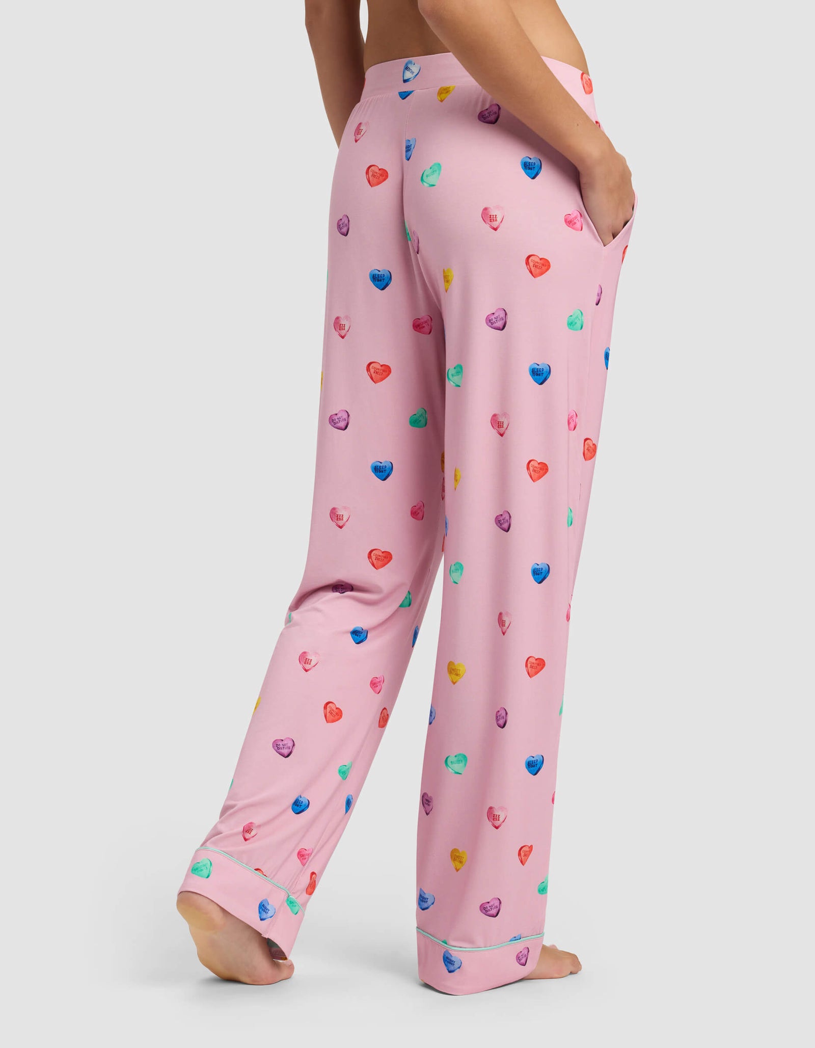 A person stands barefoot from behind wearing Cozy Earth's Women's Bamboo Stretch-Knit Pajama Pant in light pink, featuring colorful, scattered heart patterns and a hand in a side pocket.