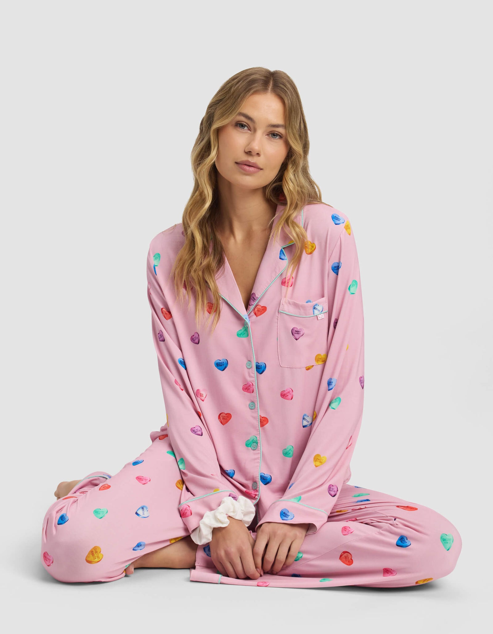 A woman with long blonde hair sits cross-legged against a plain light background, wearing Cozy Earth's Women's Bamboo Stretch-Knit Pajama Pant in pink with colorful heart patterns.