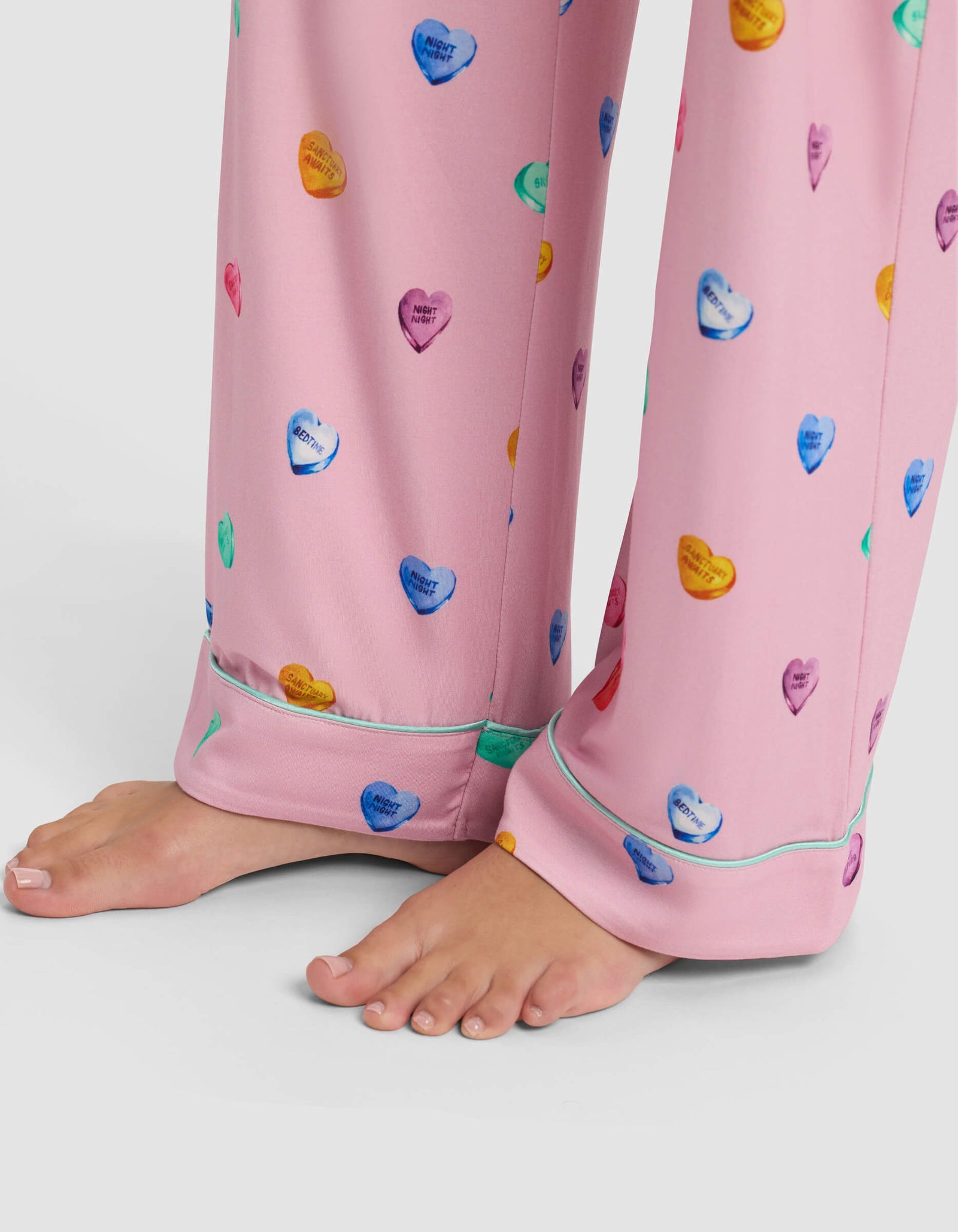 Bare feet on a white surface, wearing Cozy Earth Women's Bamboo Stretch-Knit Pajama Pant in pink, featuring colorful candy hearts and small printed messages.