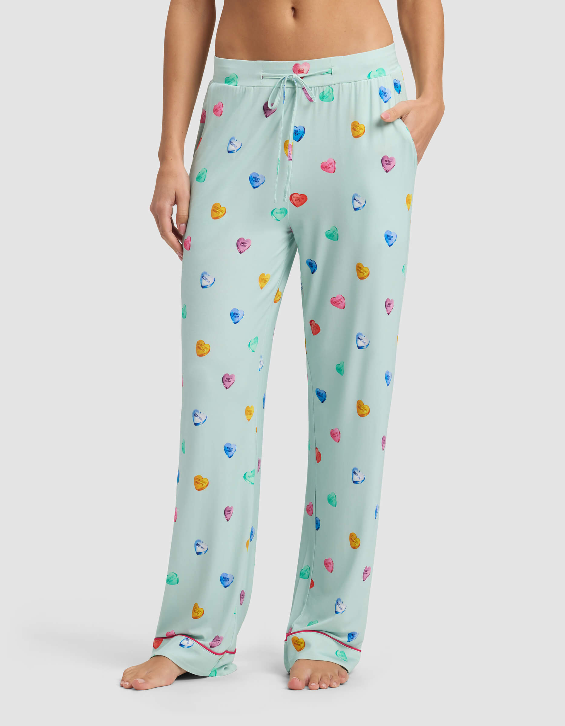 A person stands barefoot wearing Cozy Earth’s Women's Bamboo Stretch-Knit Pajama Pant in light blue with colorful heart-shaped candy prints, featuring a drawstring waist and pockets, with one hand resting in a pocket.|Color:Seafoam Sleepy Hearts