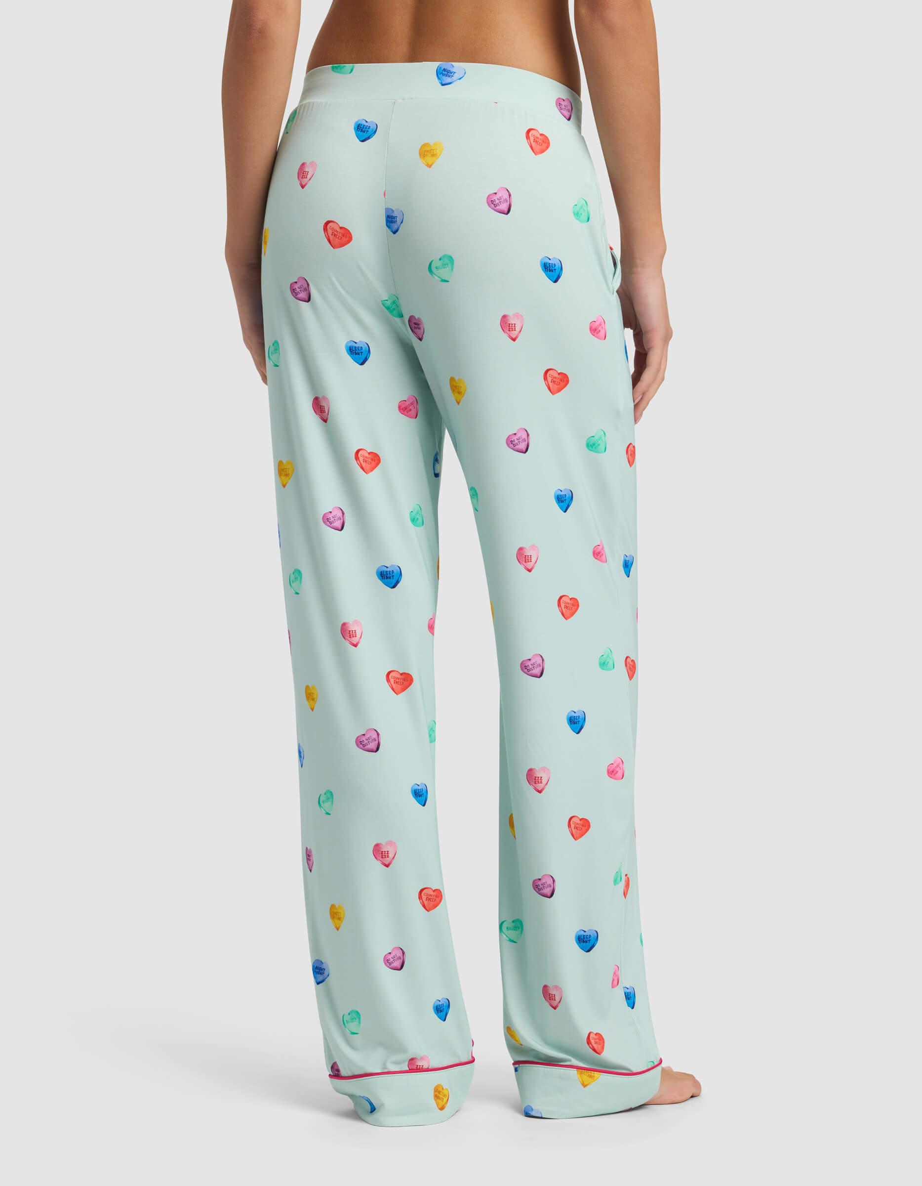 A person stands barefoot, facing away, wearing Cozy Earth Women's Bamboo Stretch-Knit Pajama Pant in light green with a colorful heart pattern on a plain white background.|Color:Seafoam Sleepy Hearts