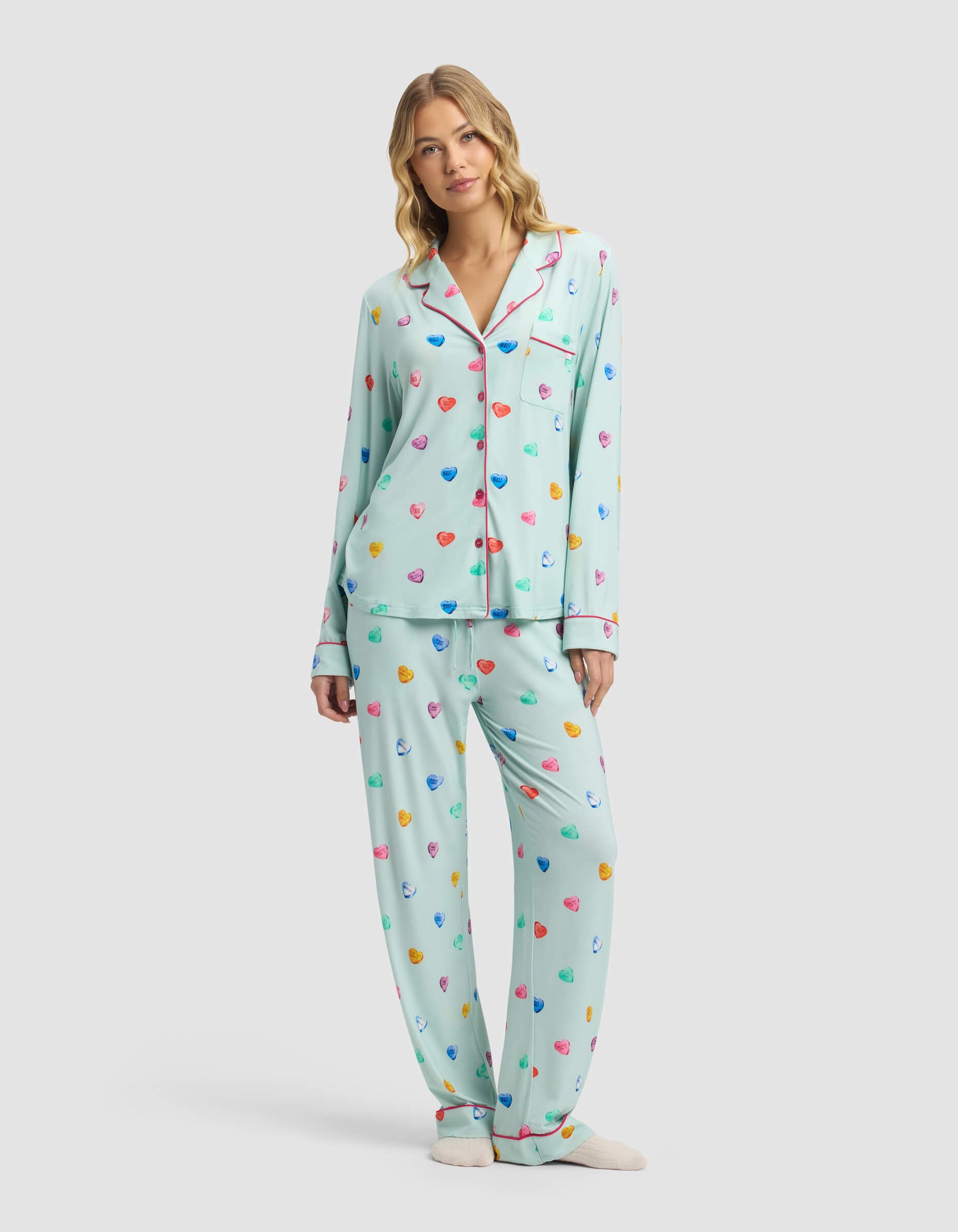A woman wears Cozy Earth's Women's Bamboo Stretch-Knit Pajama Pant in light blue with a colorful heart pattern, button-up top, and cream slippers, standing against a plain light background.