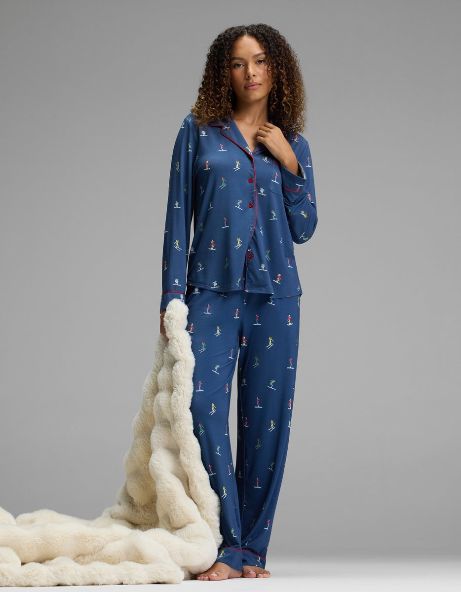A woman wearing navy blue Cozy Earth Women’s Bamboo Stretch-Knit Pajama Pant and a matching button-down top with a colorful skier print and red piping, holding a cream-colored faux fur throw blanket.