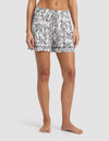 A woman wears Cozy Earth's Women's Bamboo Stretch-Knit Pajama Shorts, featuring a black floral pattern and drawstring waistband, standing barefoot against a plain light background.|Color:Jet Black Toile