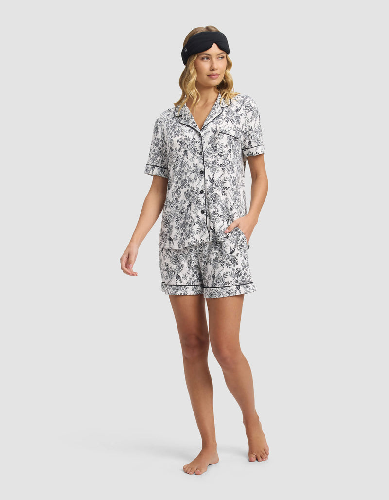 A woman stands barefoot against a plain light background, wearing Cozy Earth's Women's Bamboo Stretch-Knit Pajama Short set in a white and black floral pattern, featuring a short-sleeve button-up top and matching shorts with a black sleep mask.