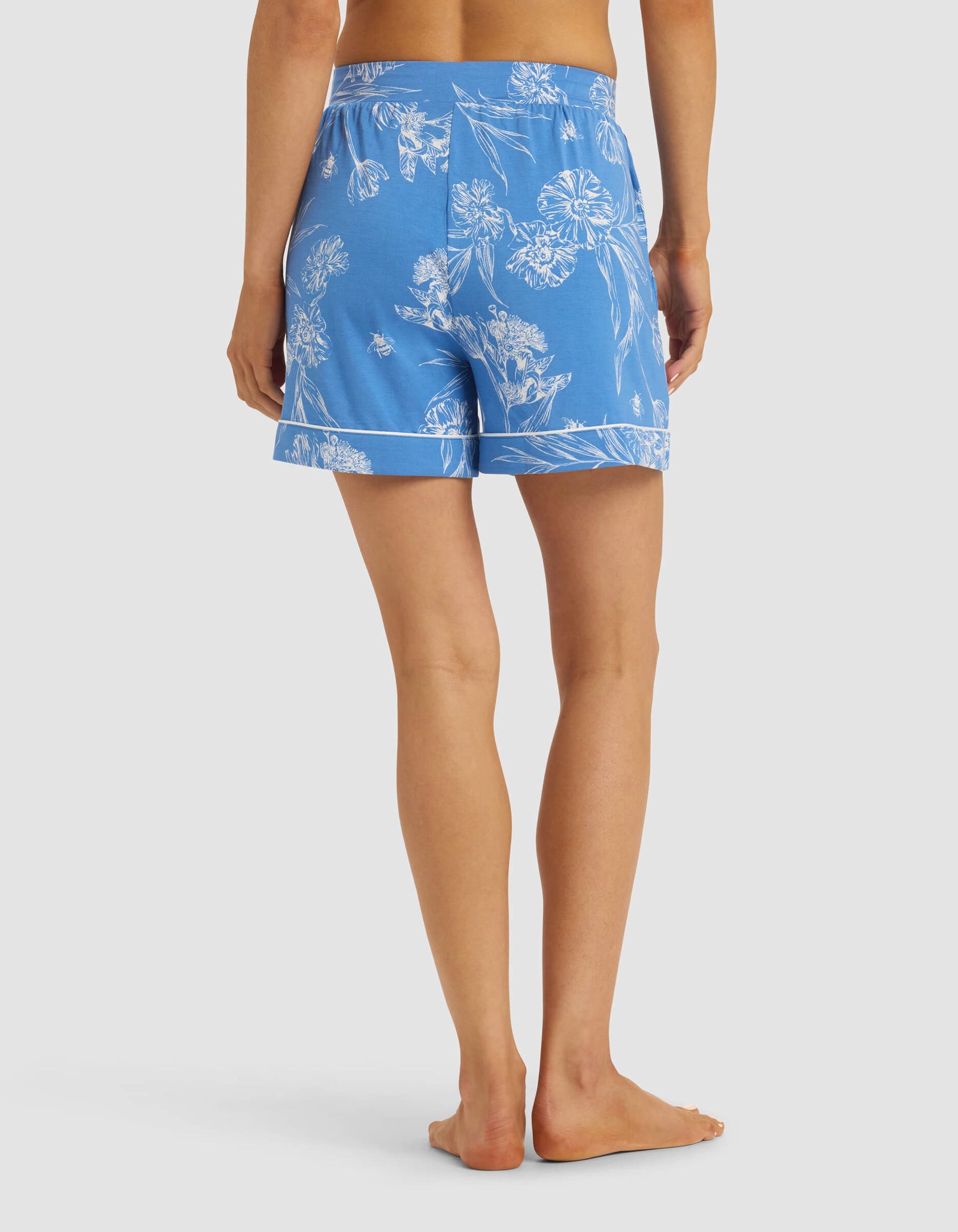 A person stands barefoot, facing away, wearing Cozy Earth Women's Bamboo Stretch-Knit Pajama Shorts in blue with a white floral pattern, featuring a relaxed fit and elastic waistband against a plain light background.