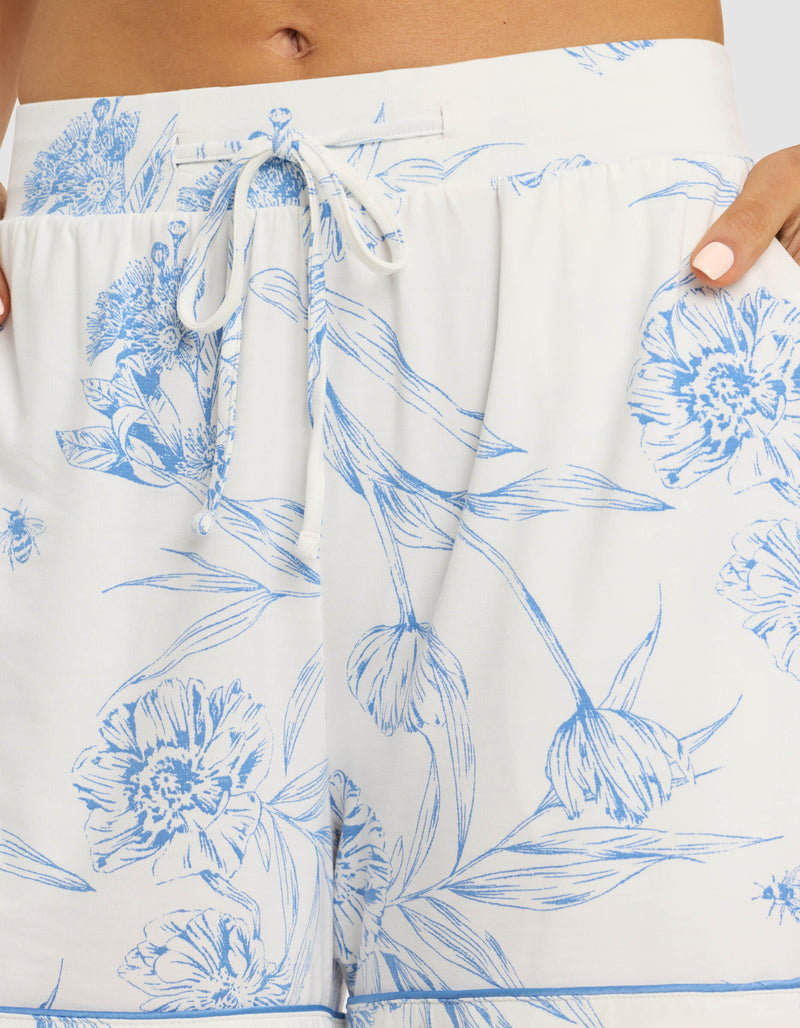 A close-up of a person wearing Cozy Earth Women's Bamboo Stretch-Knit Pajama Shorts in white with a blue floral pattern and drawstring waistband, hand resting on their hip.