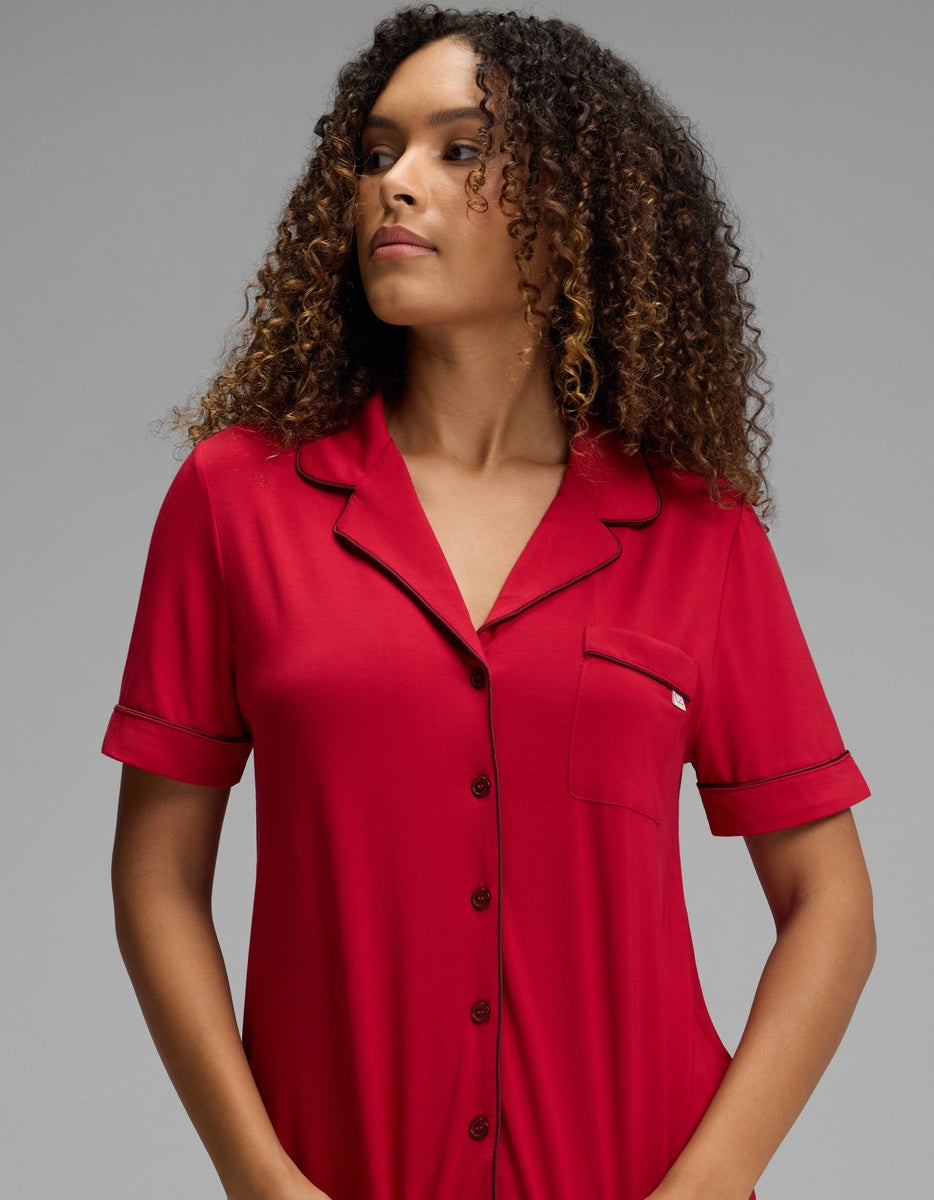 A woman wearing a Cozy Earth Women’s Bamboo Stretch-Knit Short Sleeve Pajama Top in red, featuring black trim, a button-down front, and a chest pocket.