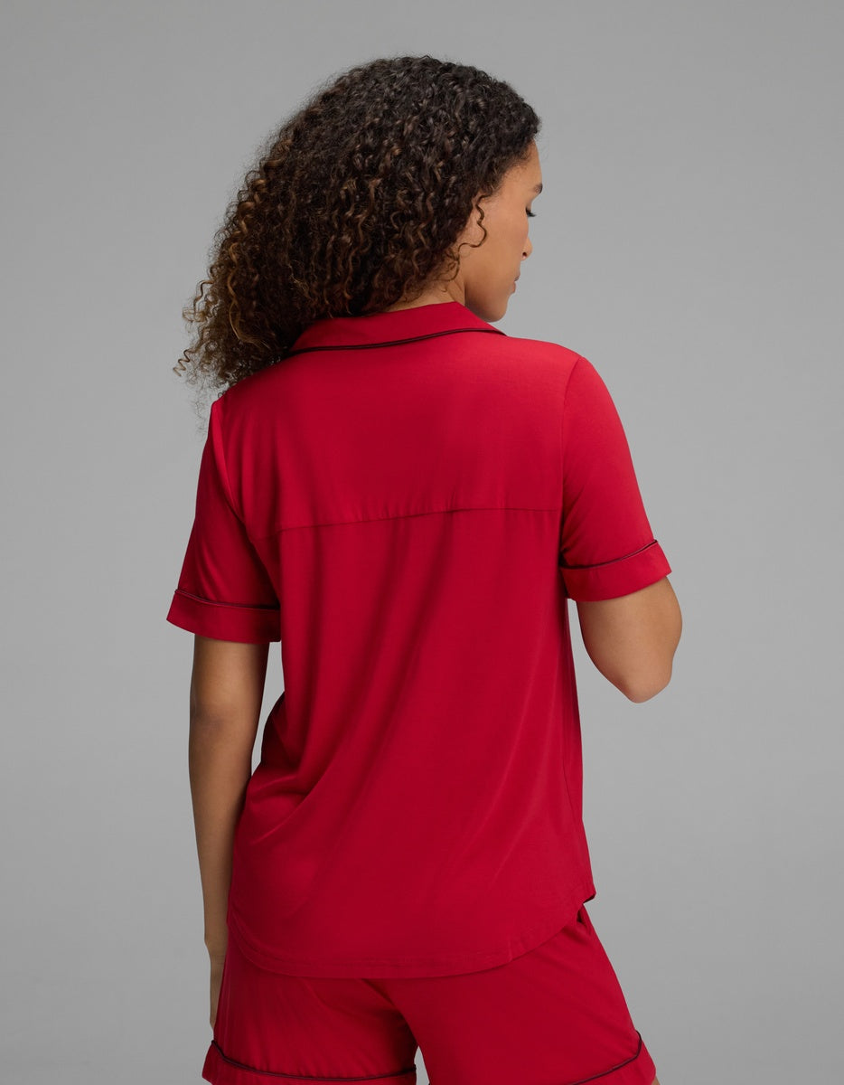 View from the back of a person wearing a red Women’s Bamboo Stretch-Knit Short Sleeve Pajama Top by Cozy Earth, featuring a collared neckline and thin black trim on the collar and sleeve cuffs.