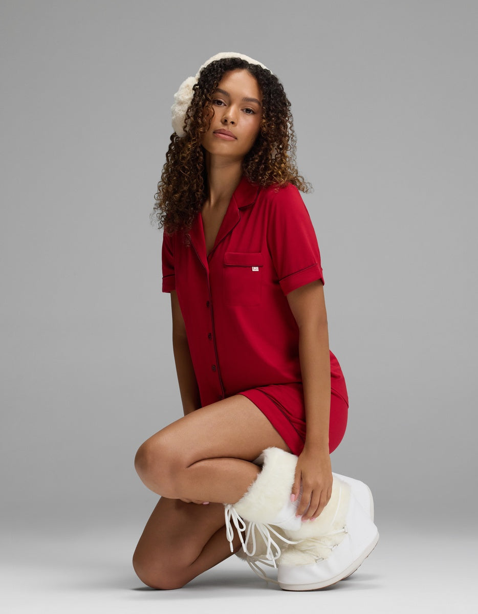 A woman wearing a red Women’s Bamboo Stretch-Knit Short Sleeve Pajama Top, white earmuffs, and white winter boots kneels against a grey background.