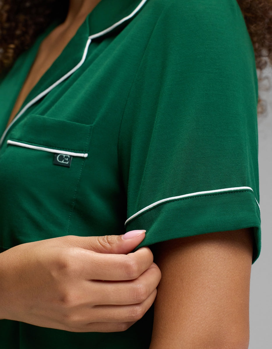 A person pulls on the hem of a short-sleeved green Cozy Earth Women’s Bamboo Stretch-Knit Short Sleeve Pajama Top featuring white piping and a small chest pocket.