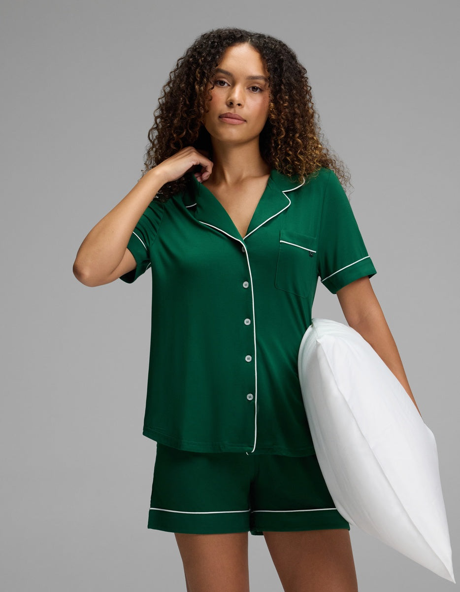 A woman wearing a dark green, short-sleeved Women’s Bamboo Stretch-Knit Short Sleeve Pajama Top and matching shorts with white piping, holding a white pillow under her arm.