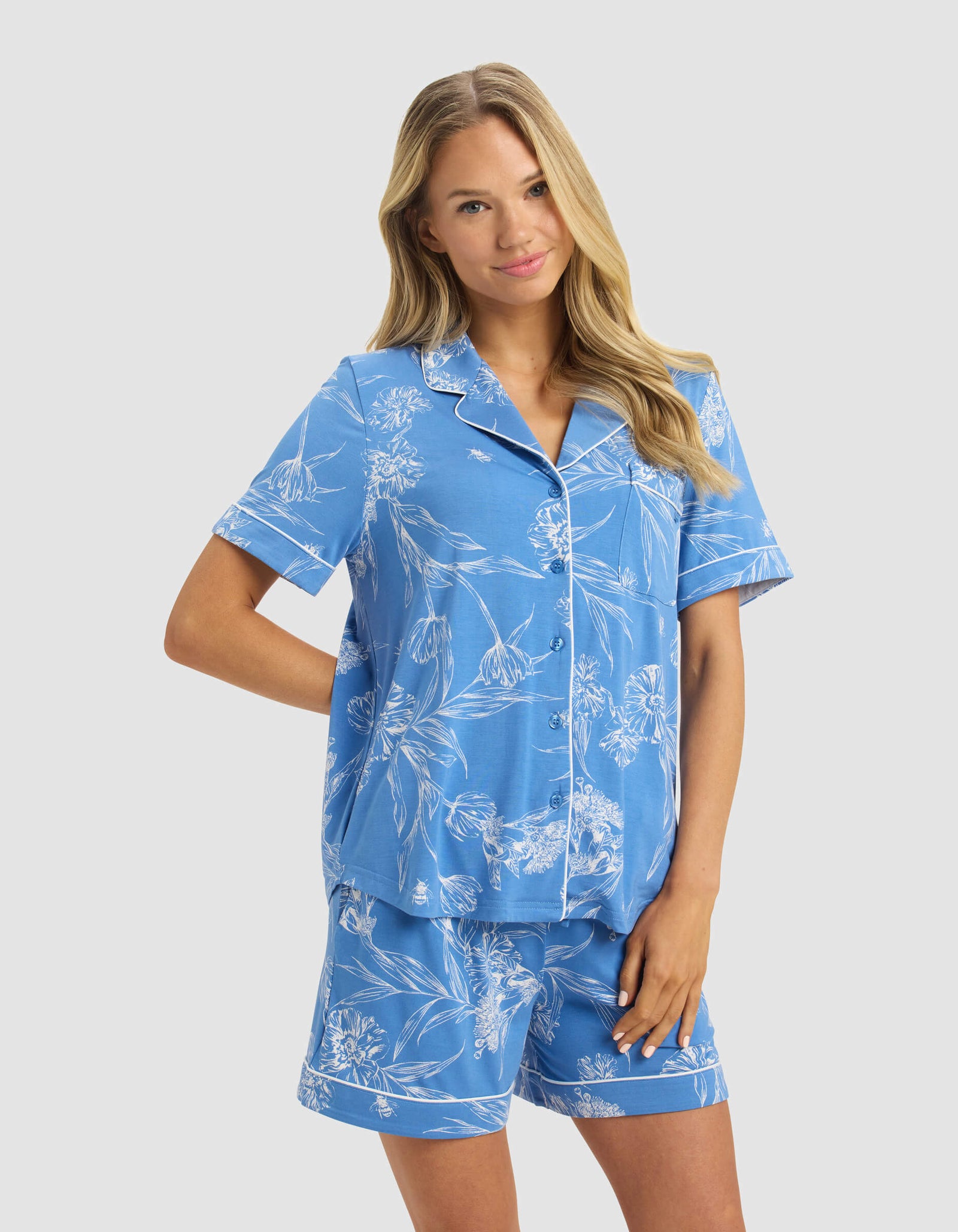 A woman with long blonde hair wears the Cozy Earth Women's Bamboo Stretch-Knit Short Sleeve Pajama Top in blue with white floral patterns, paired with matching shorts, standing and smiling against a plain light gray background.