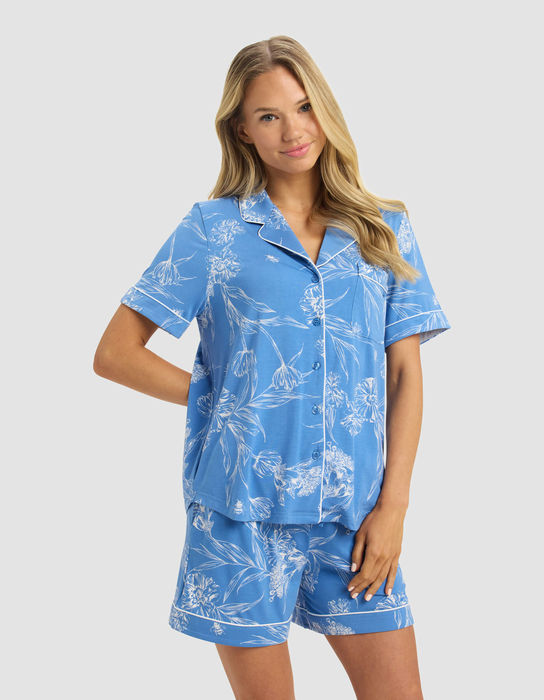A woman with long blonde hair wears the Cozy Earth Women's Bamboo Stretch-Knit Short Sleeve Pajama Top in blue with white floral patterns, paired with matching shorts, standing and smiling against a plain light gray background.|Color:Garden Toile in Sailor