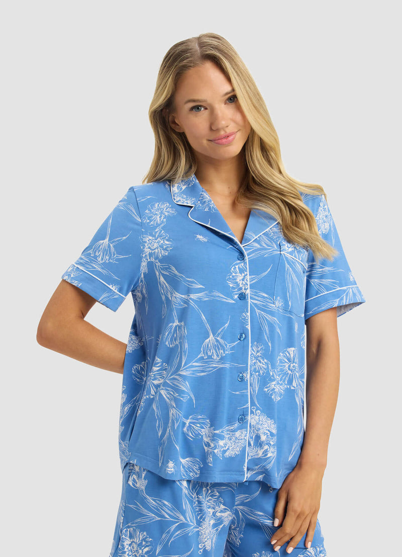 A woman with long blonde hair models the Cozy Earth Women's Bamboo Stretch-Knit Short Sleeve & Pant Pajama Set in light blue with white floral prints and piping, standing against a light gray background and smiling softly with one hand on her hip.