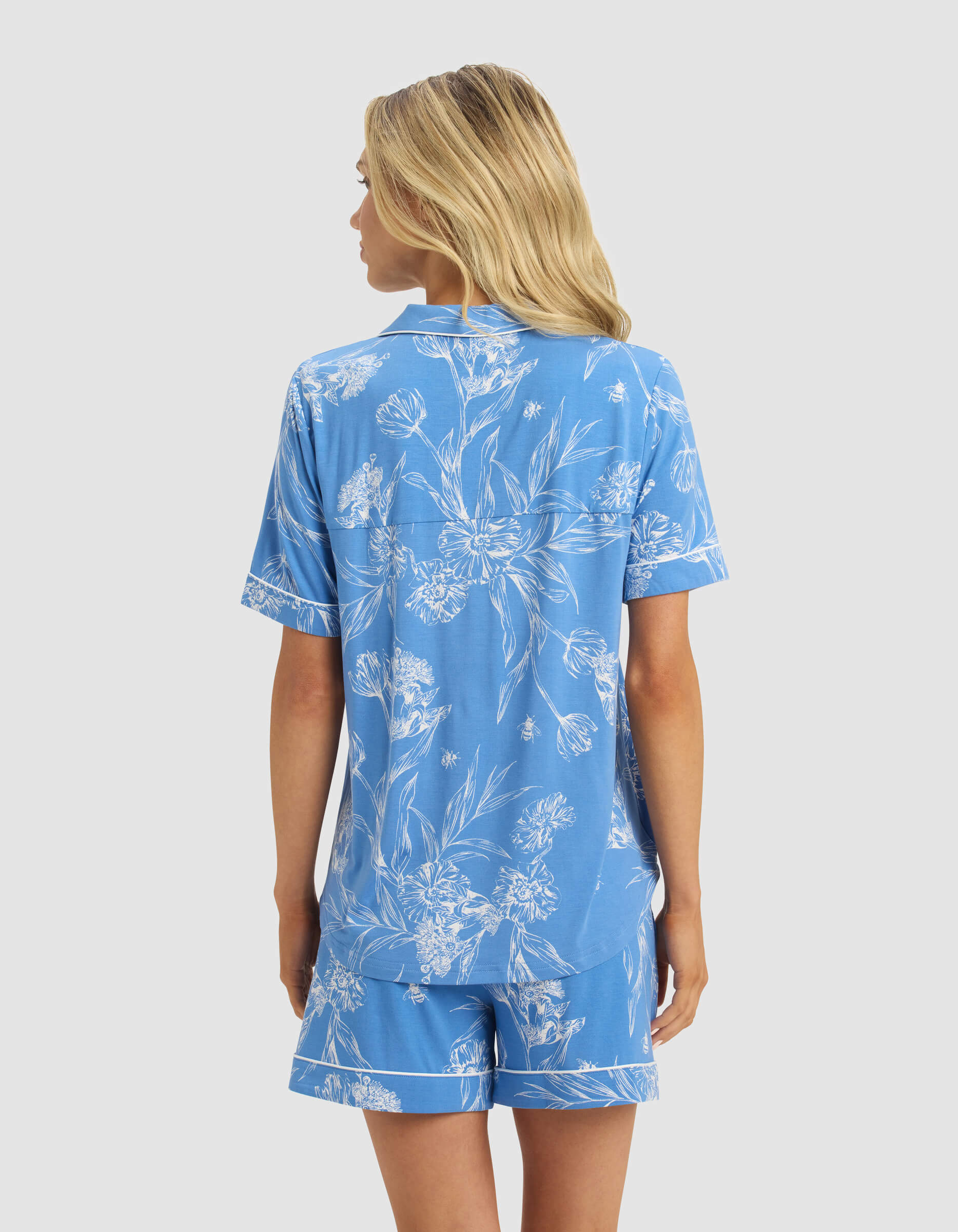 A woman with long blonde hair is seen from the back wearing a Cozy Earth Women's Bamboo Stretch-Knit Short Sleeve Pajama Top in blue with white floral patterns, set against a plain light gray background.|Color:Garden Toile in Sailor