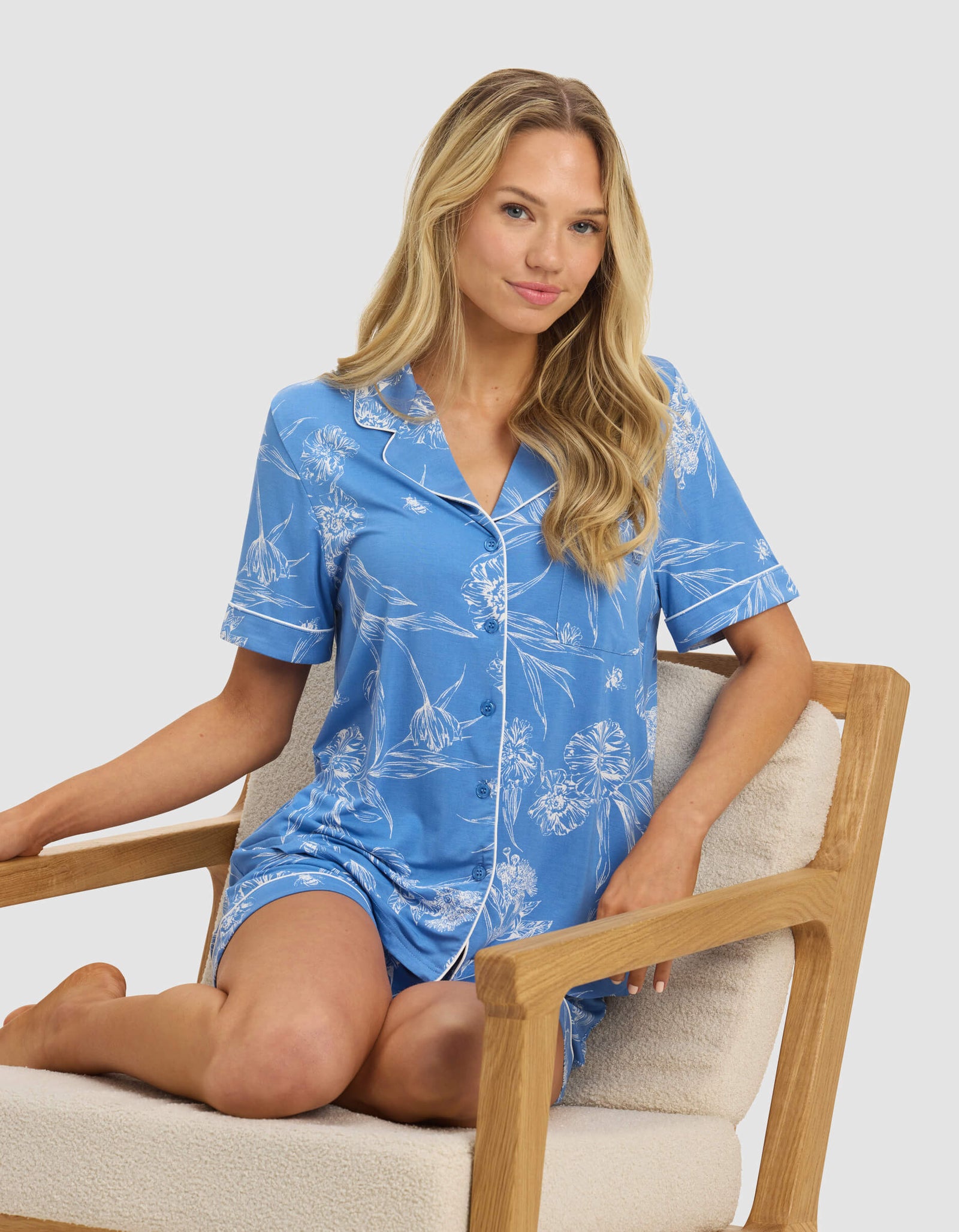 A woman with long blonde hair sits on a wooden chair, wearing Cozy Earth's Women's Bamboo Stretch-Knit Short Sleeve Pajama Top, light blue with a white floral pattern and piping, against a plain light background.