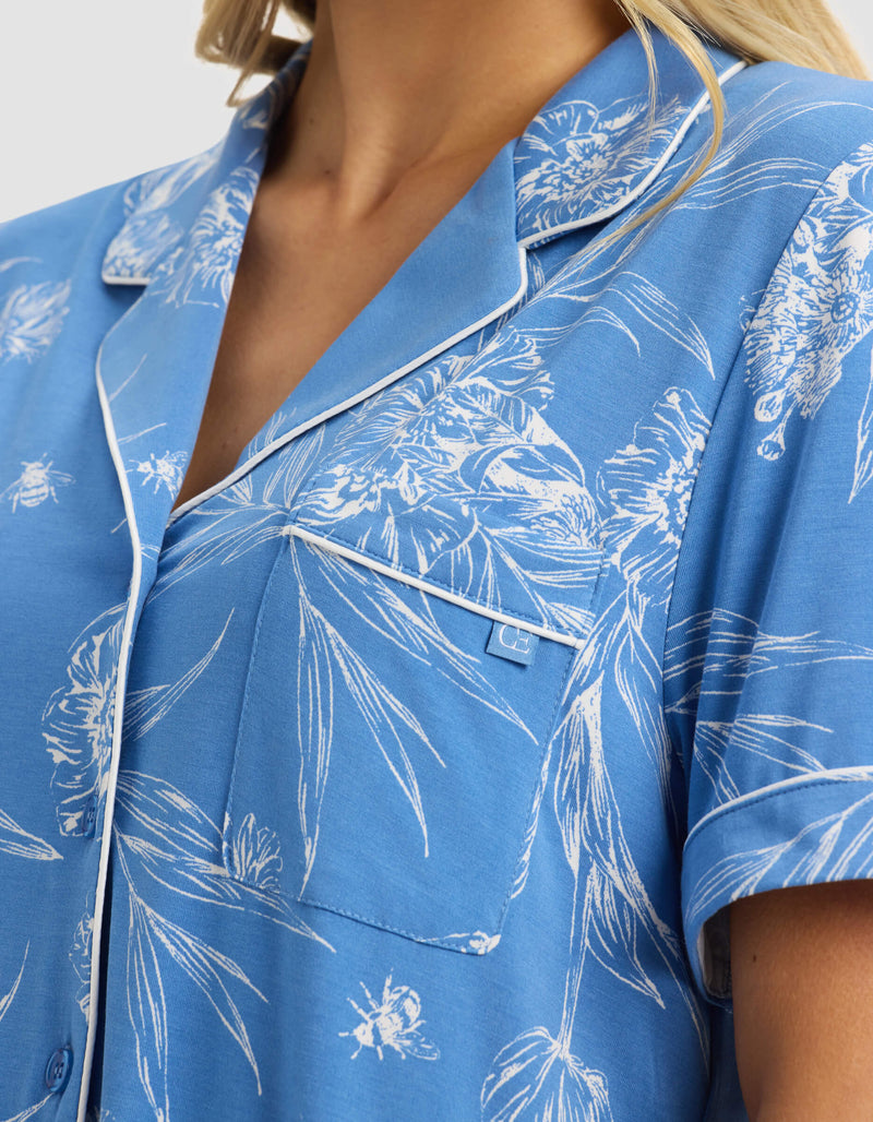 A close-up of a woman wearing the Cozy Earth Women's Bamboo Stretch-Knit Short Sleeve Pajama Top in blue with a white floral and bee print, featuring white piping on the collar, lapel, and pocket.