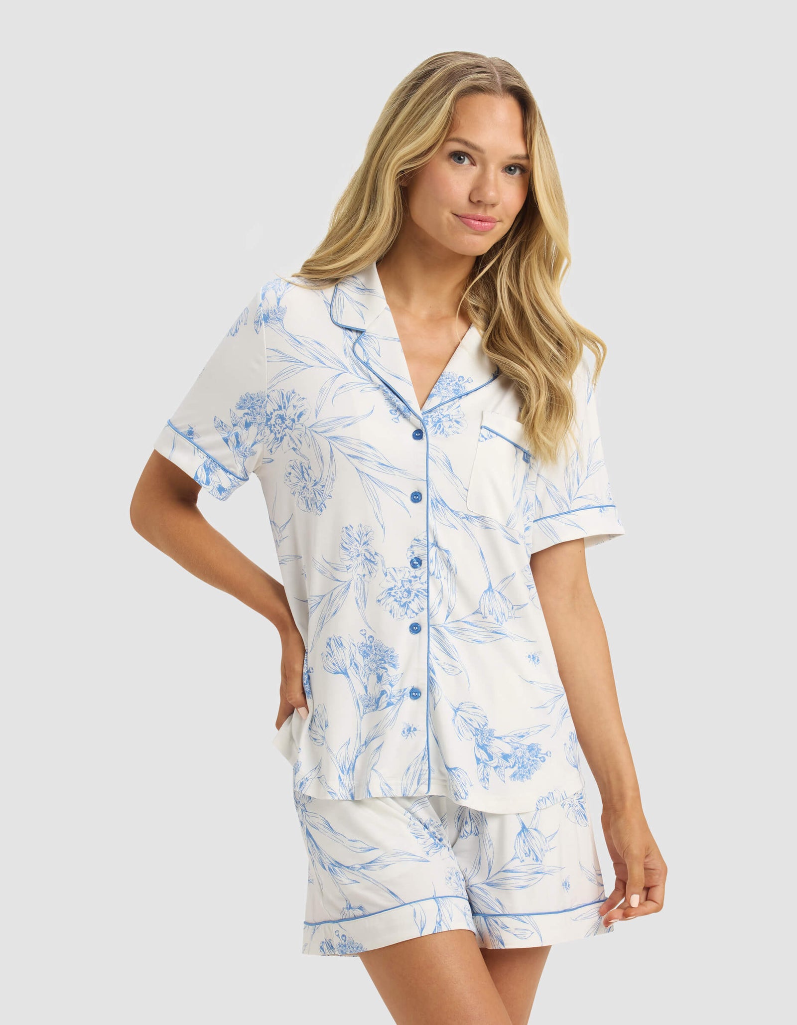 A woman with long blonde hair wears the Cozy Earth Women's Bamboo Stretch-Knit Short Sleeve Pajama Top, featuring a collar, front buttons, chest pocket, and blue floral patterns on white. She stands against a light gray background.