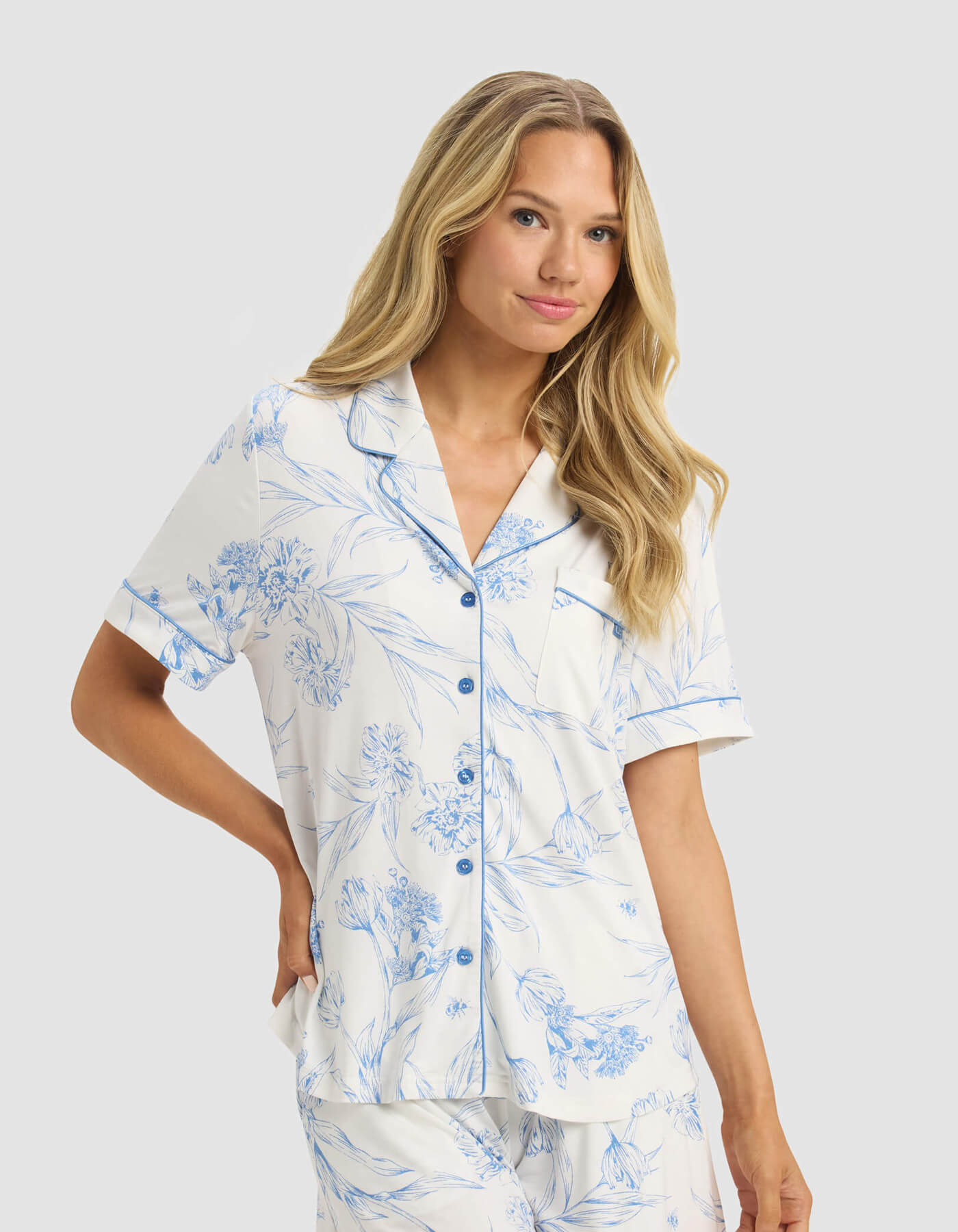 A woman with long blonde hair models the Cozy Earth Women's Bamboo Stretch-Knit Short Sleeve & Pant Pajama Set in white with a blue floral pattern, standing against a plain light gray background.