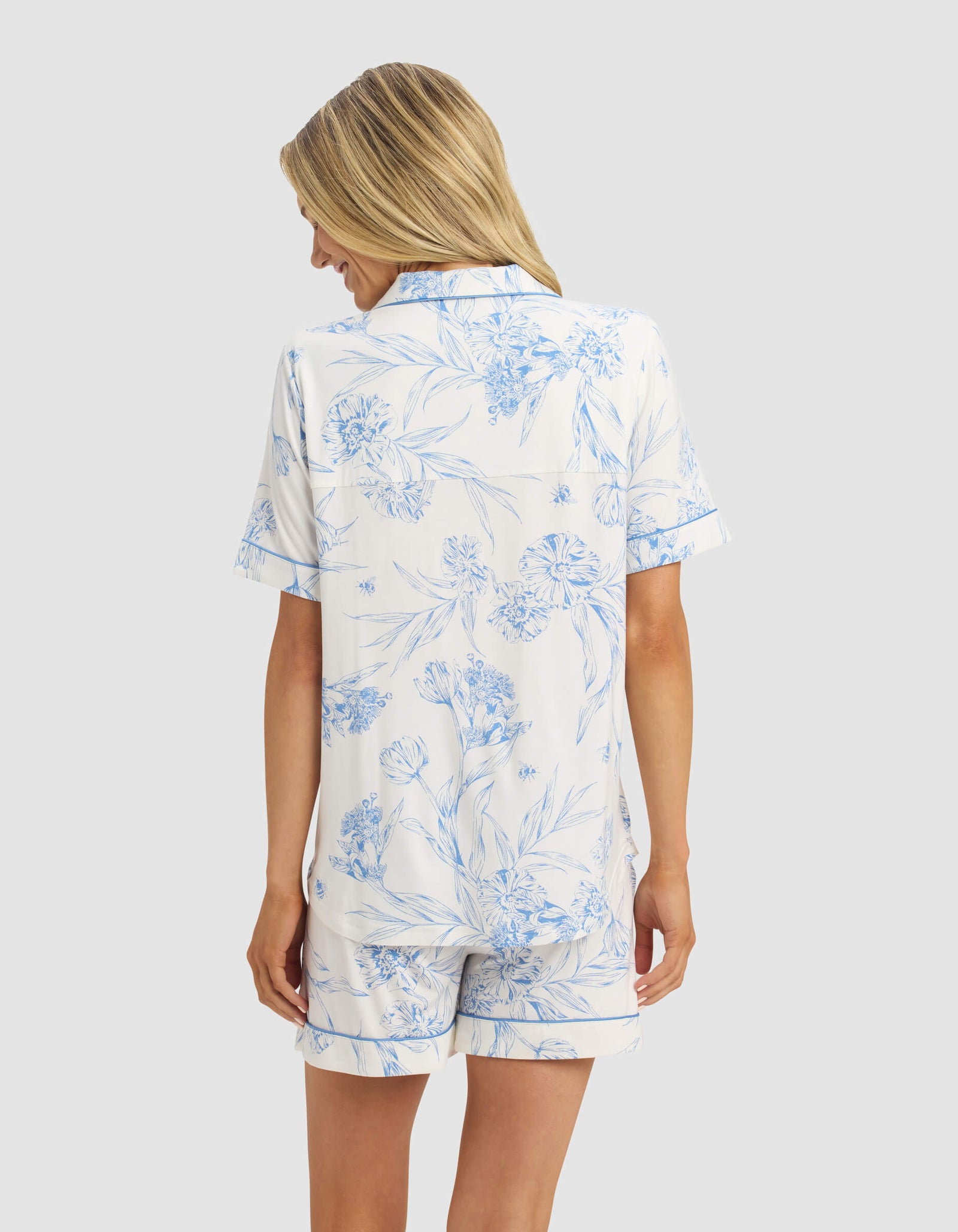 A woman with long blonde hair is seen from behind, wearing the Cozy Earth Women's Bamboo Stretch-Knit Short Sleeve Pajama Top and matching shorts in a light blue floral pattern on a white background.