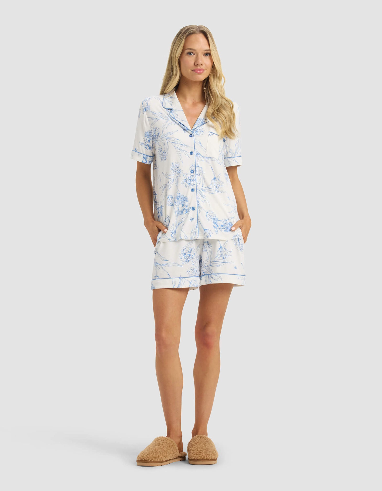 A woman stands facing forward wearing a Cozy Earth Women's Bamboo Stretch-Knit Short Sleeve Pajama Top in light blue and white floral, matching shorts, and tan fuzzy slippers against a plain light background.