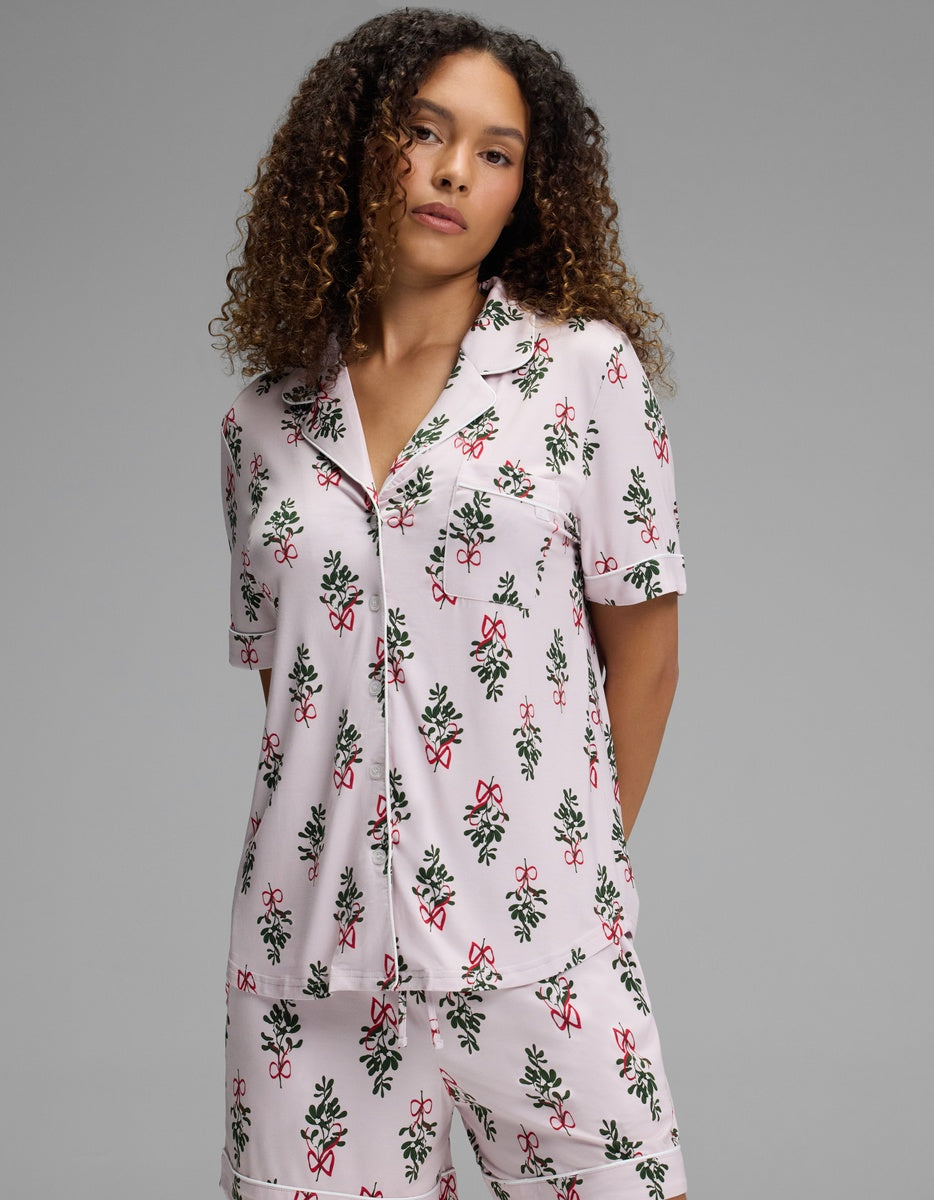 A woman wearing a Cozy Earth Women’s Bamboo Stretch-Knit Short Sleeve Pajama Top with a pink background and a holiday-themed pattern of bows and holly.