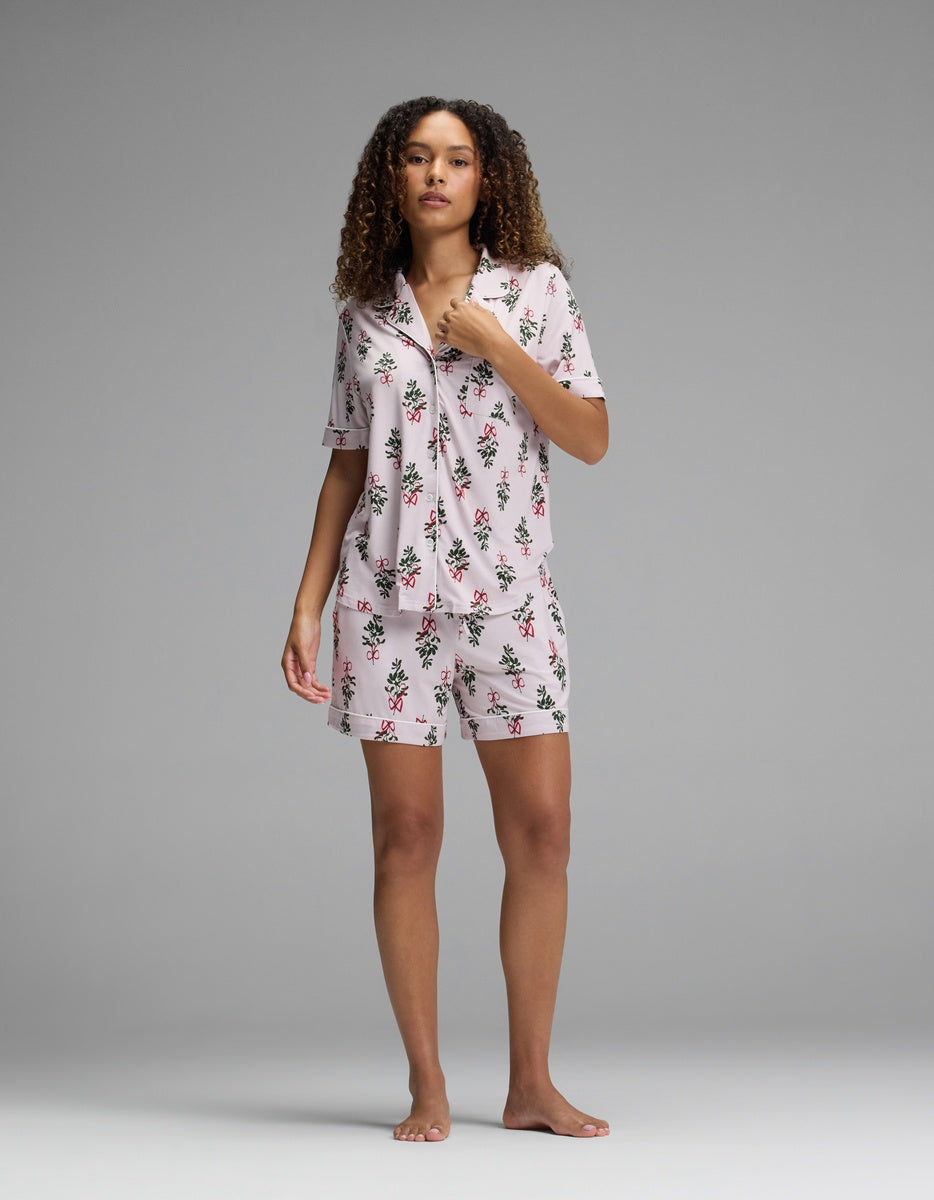 A woman wearing a Cozy Earth Women’s Bamboo Stretch-Knit Short Sleeve Pajama Top and matching shorts in pale pink with a pattern of red bows and green sprigs.