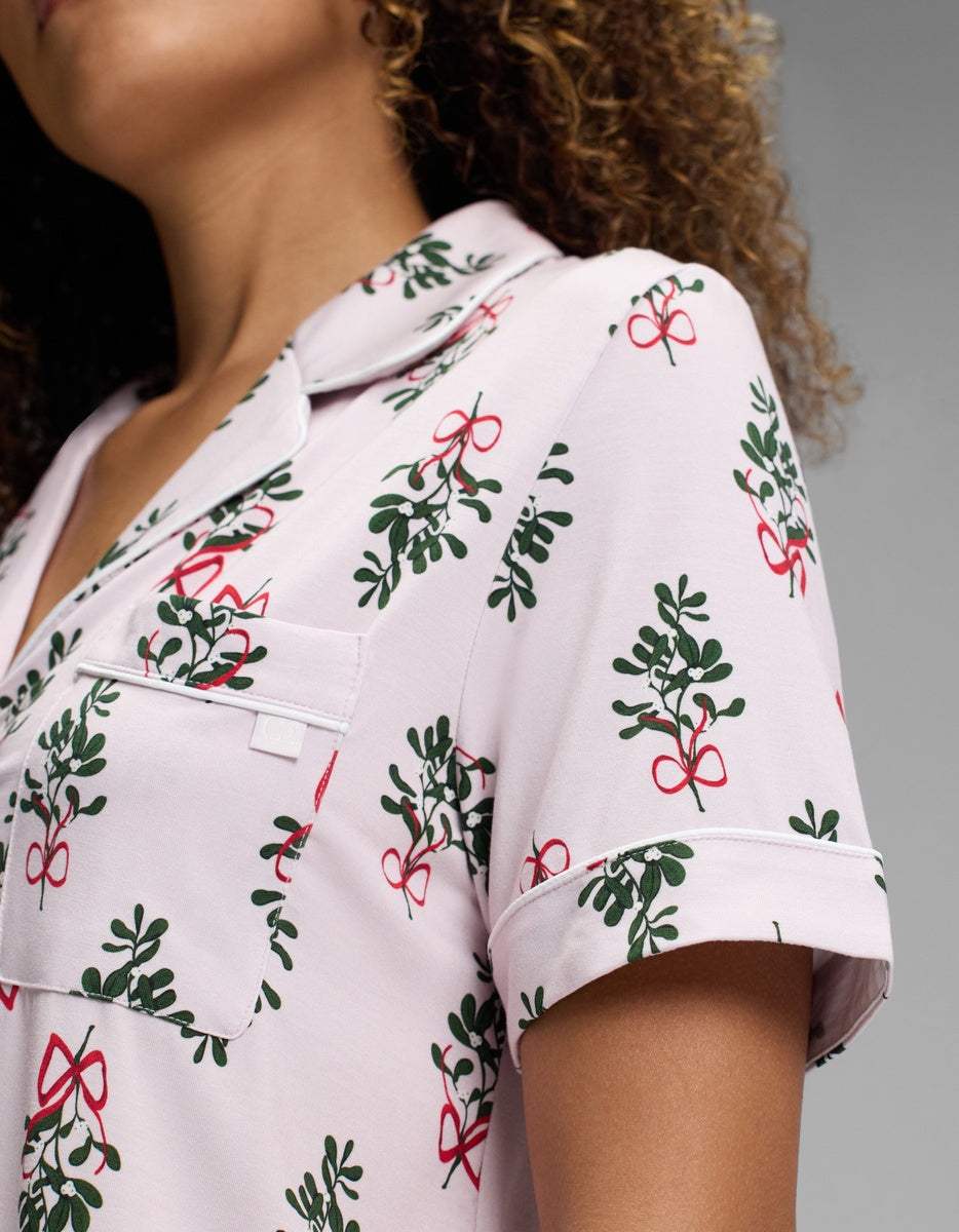 A person wearing a light pink Cozy Earth Women’s Bamboo Stretch-Knit Short Sleeve Pajama Top with a mistletoe and red ribbon print, featuring a notched collar and chest pocket.