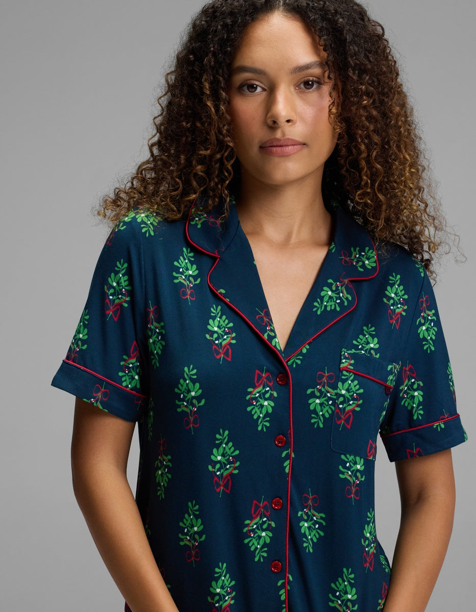 A person wears a Cozy Earth Women’s Bamboo Stretch-Knit Short Sleeve Pajama Top featuring a dark blue pattern of green mistletoe sprigs tied with red bows, finished with red piping along the collar and pocket.