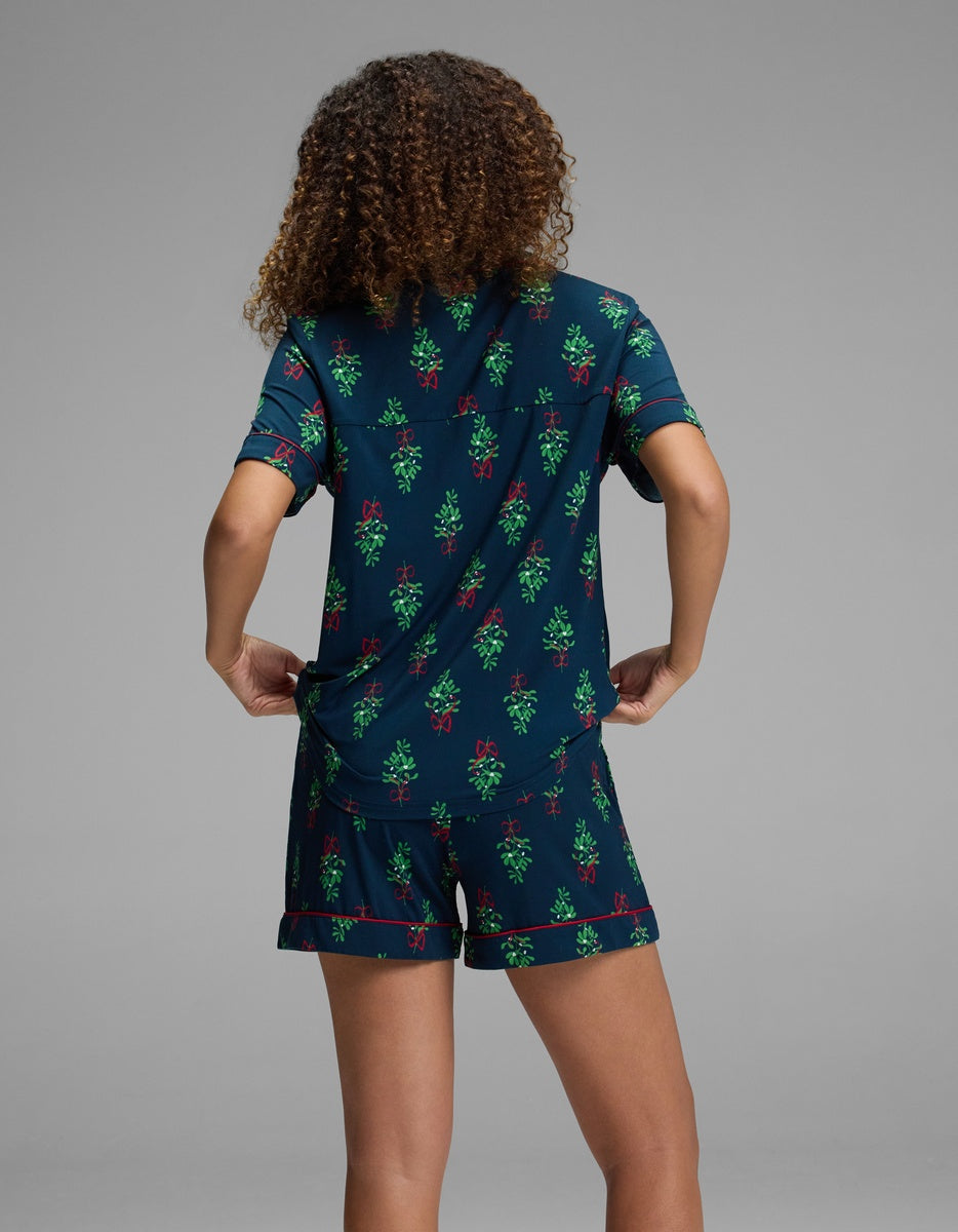 A person from behind wearing a dark blue Women’s Bamboo Stretch-Knit Short Sleeve Pajama Top and matching shorts featuring a red-and-green mistletoe print with red piping.